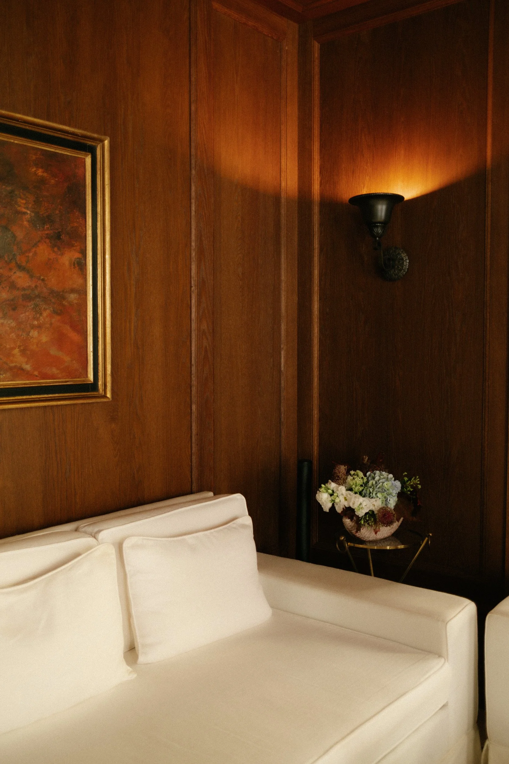A white bed with pillows against a wood-paneled wall. To the right, a vase filled with pink, purple, and white flowers is placed on a small round table, next to a black wall sconce.