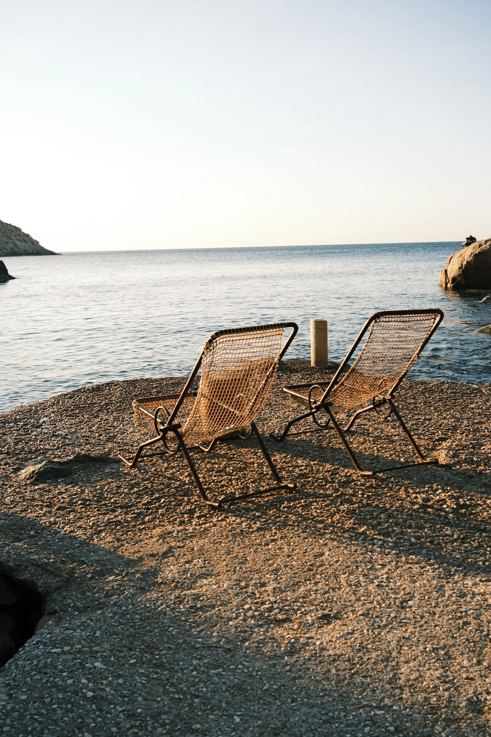 Two empty lounge chairs on a rocky beach facing the ocean with rocks and a small boat in the distance.