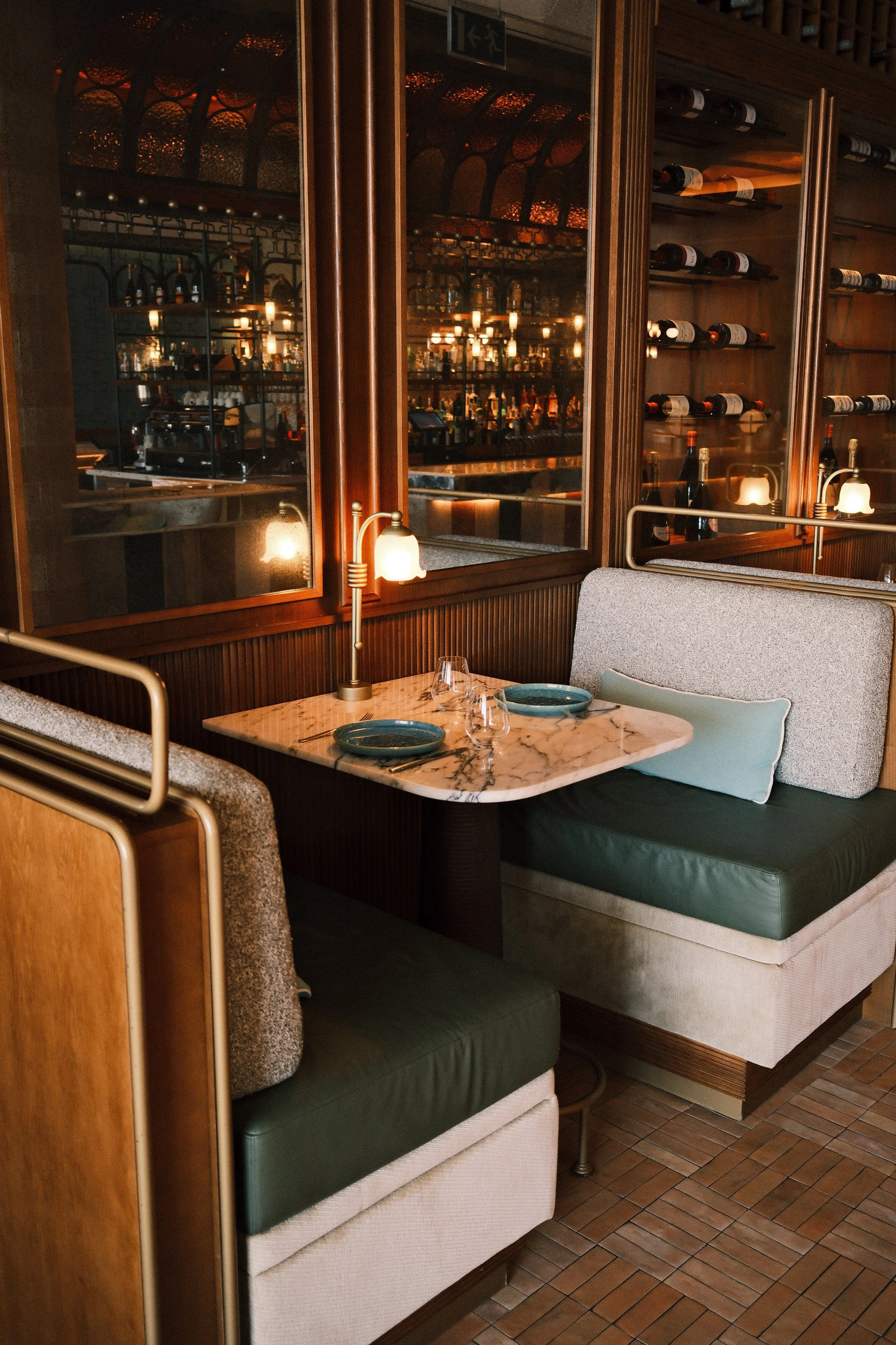 A restaurant booth with a marble table set with two green plates, wine glasses, and a napkin. There is a beige cushioned seat with a turquoise pillow and a dark green seat cushion. The booth is separated by a wood-paneled wall with a mirror, and a wall-mounted lamp provides warm lighting. In the background, there are glass shelves with wine bottles and a bar area with warm lighting and hanging lamps.