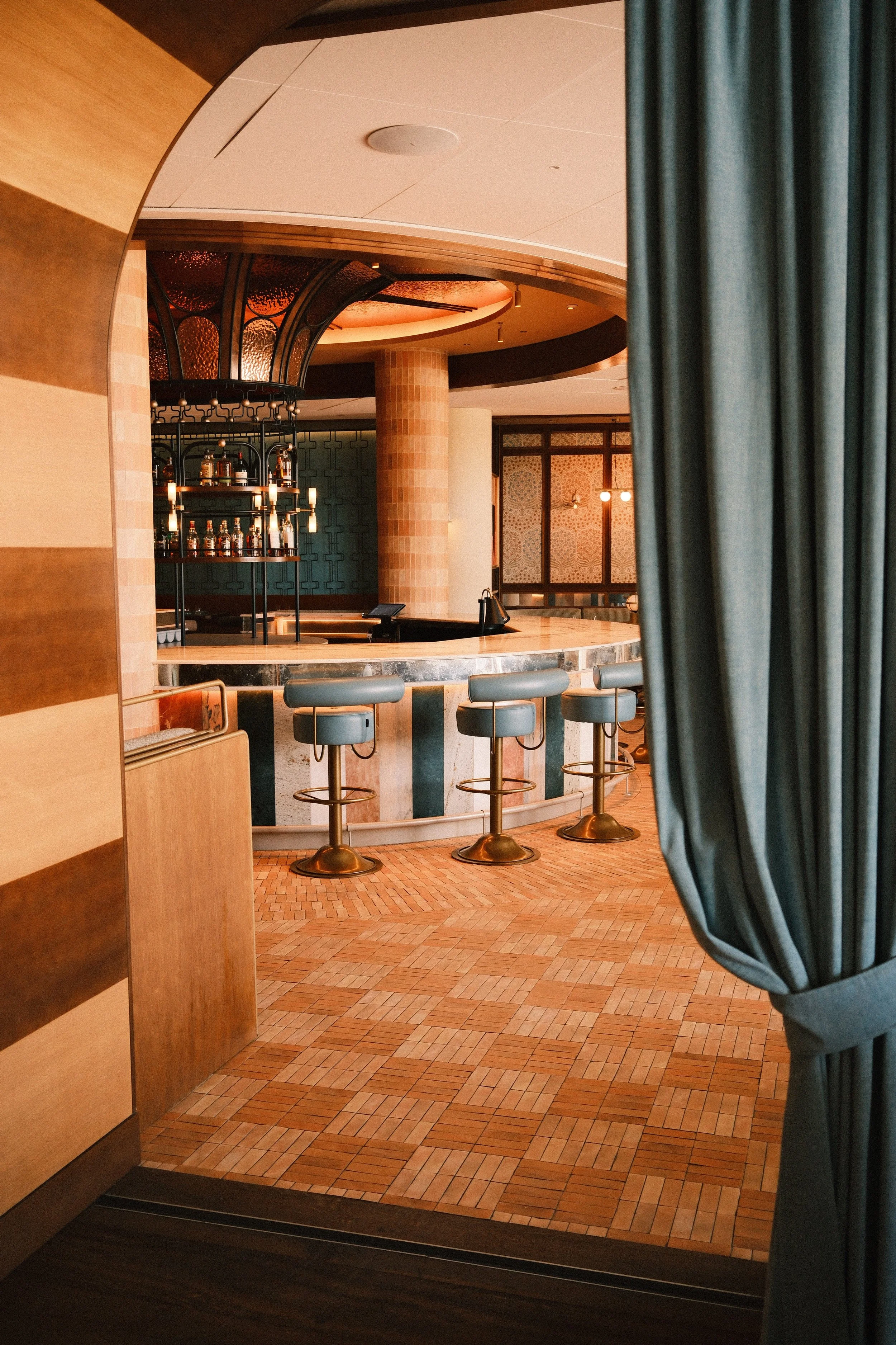 A view of an elegant bar area seen through a partially opened curtain. The bar features a rounded marble counter, gold stools, and a backdrop with a unique lighting fixture and shelves with liquor bottles. The room has warm, ambient lighting and deco