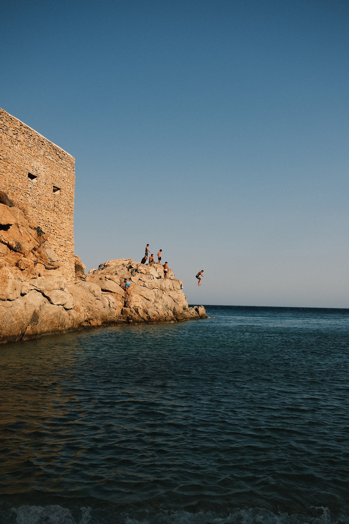 People jumping off a rocky cliff into the water at sunset, with a stone building on the left and a clear sky overhead.