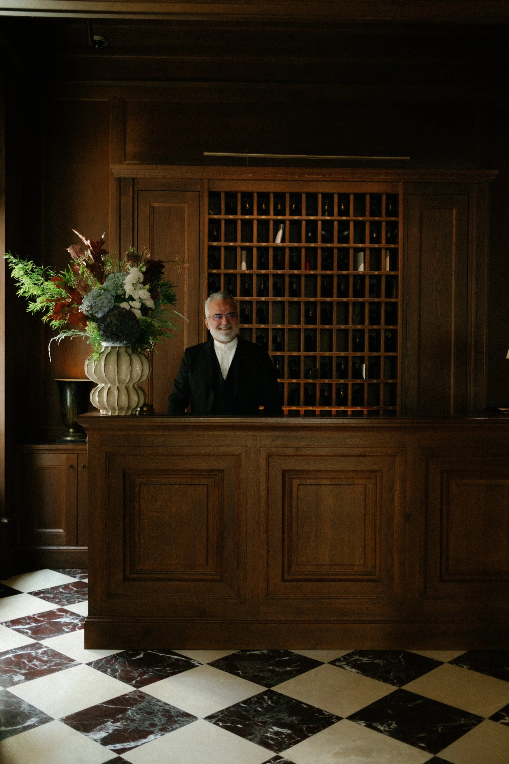 A man with white hair and a beard wearing glasses, standing behind a dark wooden reception desk, with a large floral arrangement to his left and a wine rack behind him in a wood-paneled room with a marble-checkered floor.