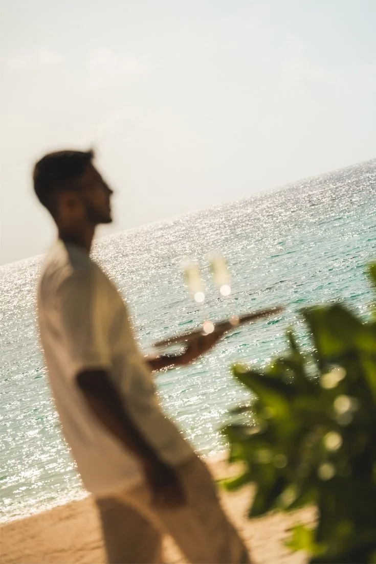 A man with dark hair and beard, wearing a light-colored shirt and shorts, holding a tray with two glasses of drink, standing on a beach near the ocean with sunlight reflecting off the water, greenery in the foreground.