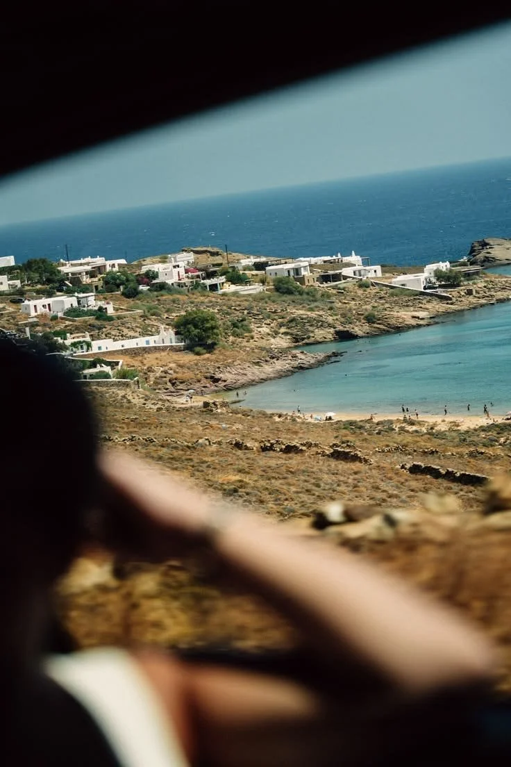 A coastal landscape viewed through a hole or gap, showing white buildings on a hillside near a beach with turquoise water and a blue ocean in the background.