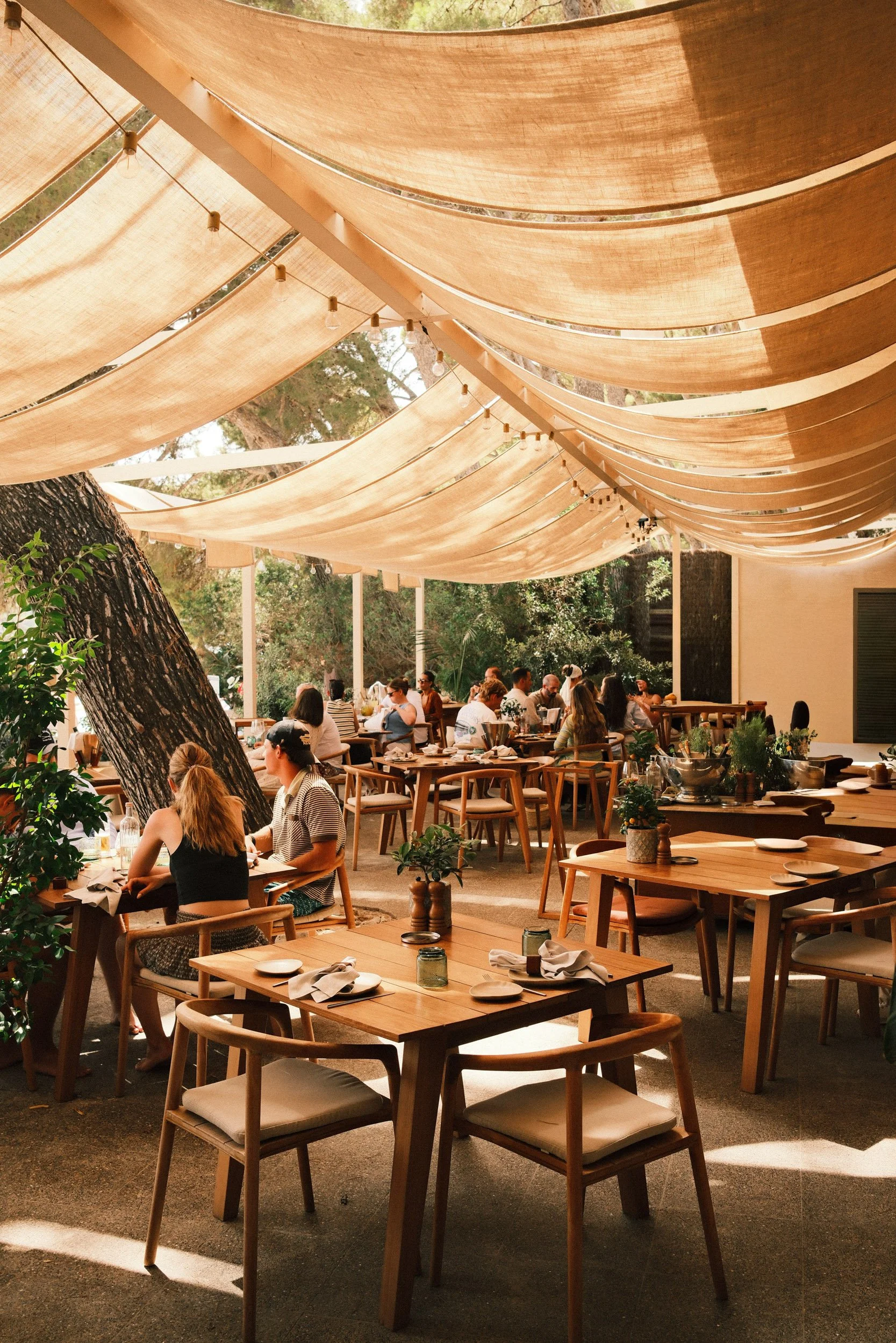 Outdoor restaurant with wooden tables and chairs, large fabric shades overhead, string lights, and patrons dining among trees and greenery.