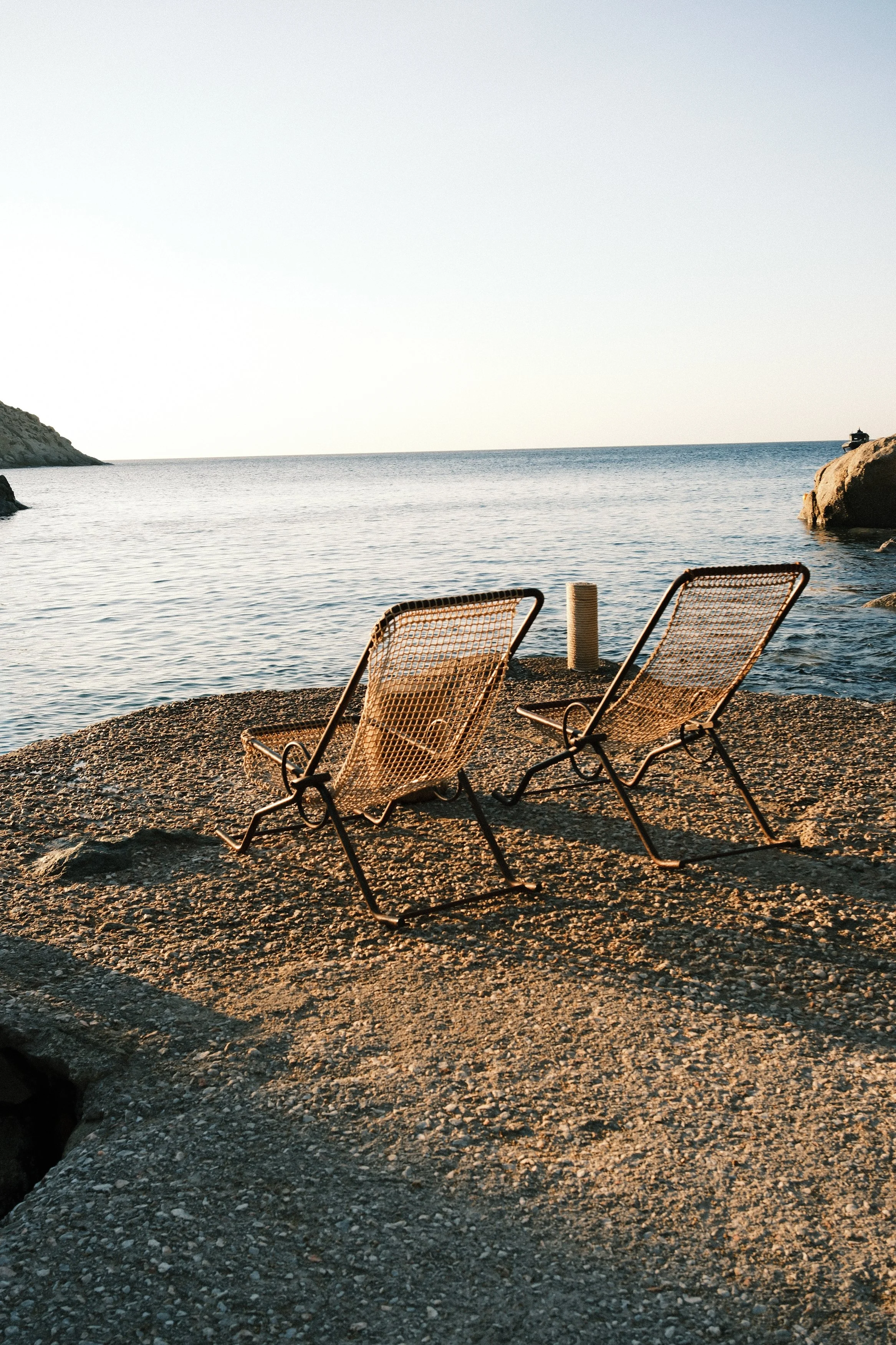 Two empty lounge chairs on a rocky beach facing the ocean, with a rocky cliff on the left and large rocks on the right, under a clear sky.