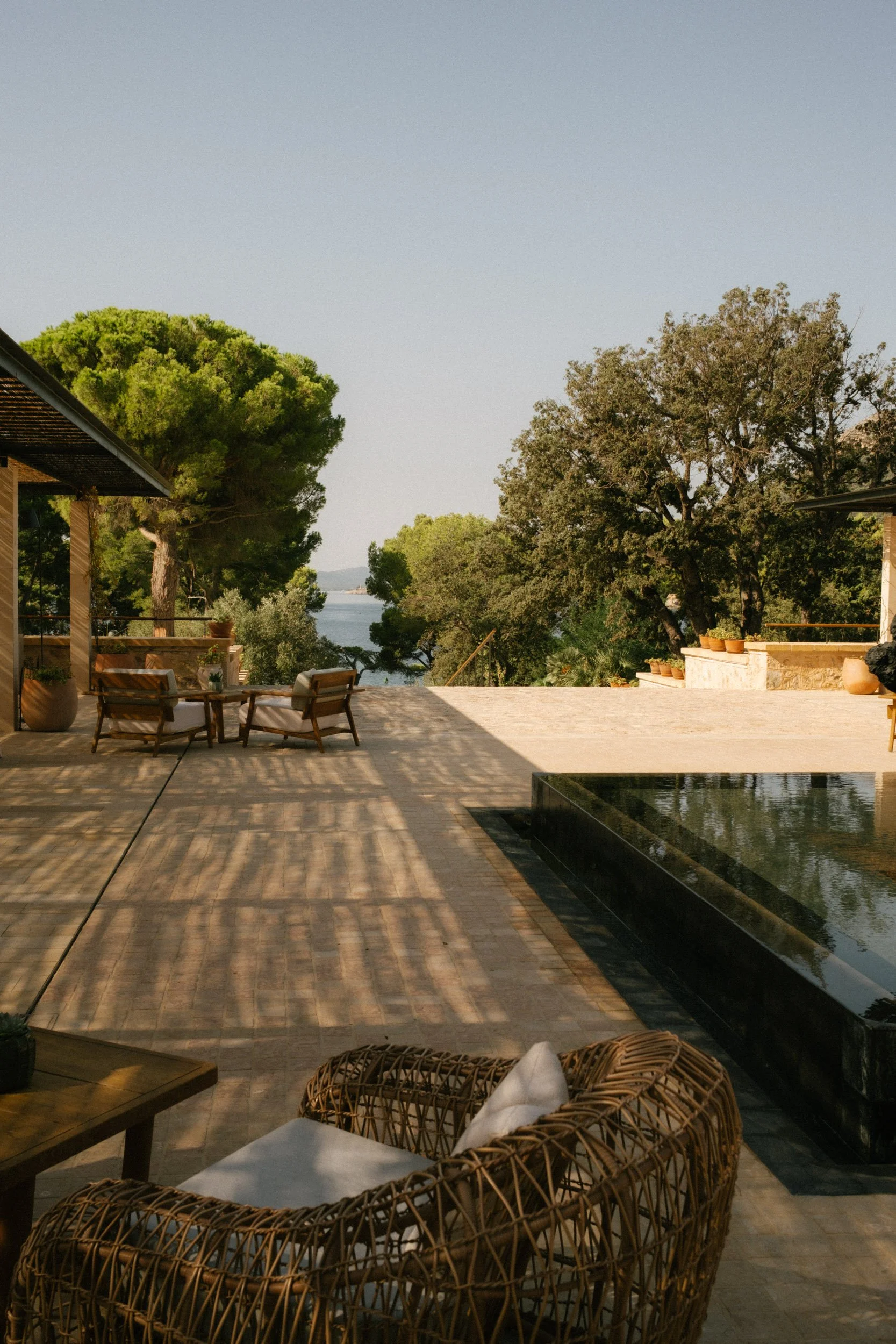 Outdoor patio with wicker chairs and a wooden table, overlooking a landscape with large trees and a body of water in the distance under a clear sky.