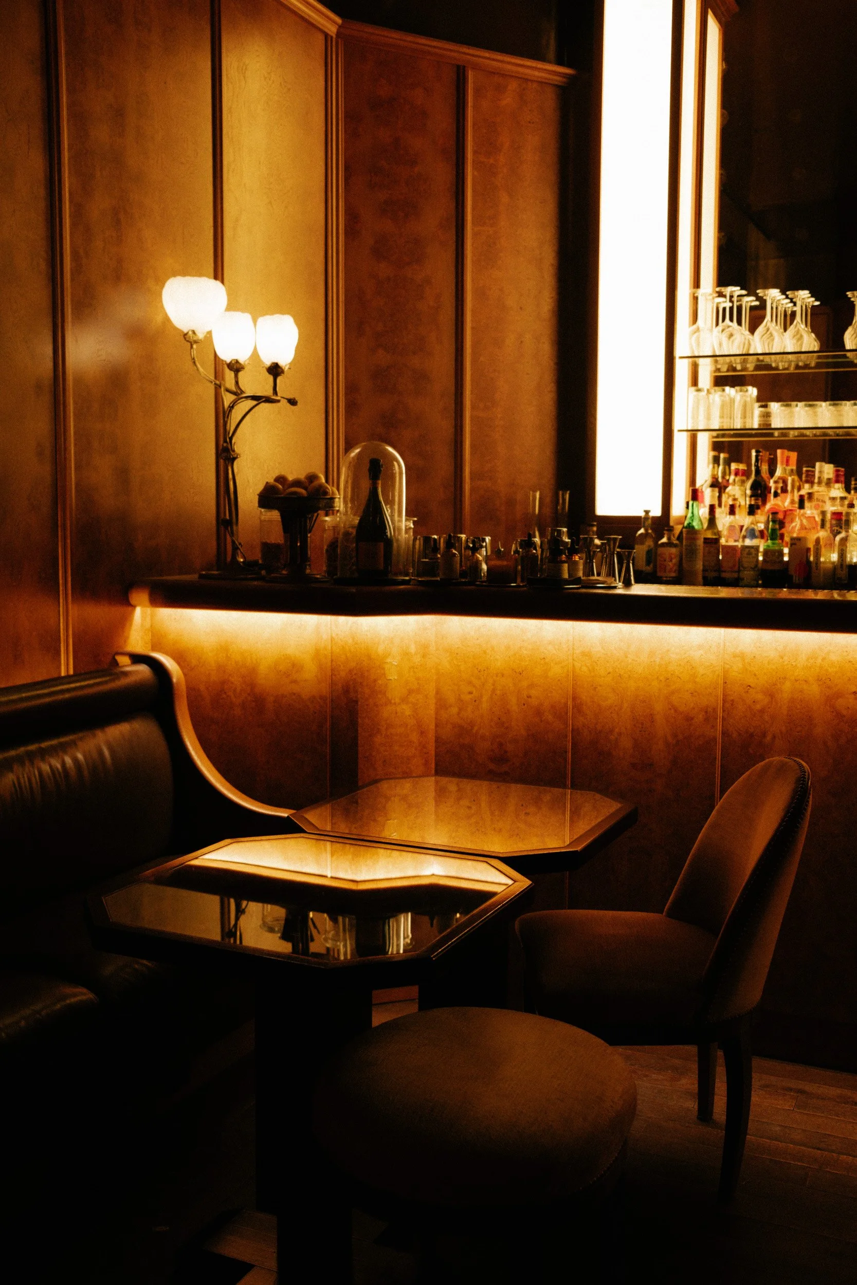 A dimly lit restaurant or lounge with wooden paneling, a bar area with various bottles and glasses, a small table with a reflective surface, and a vintage-style lamp with three frosted glass shades.
