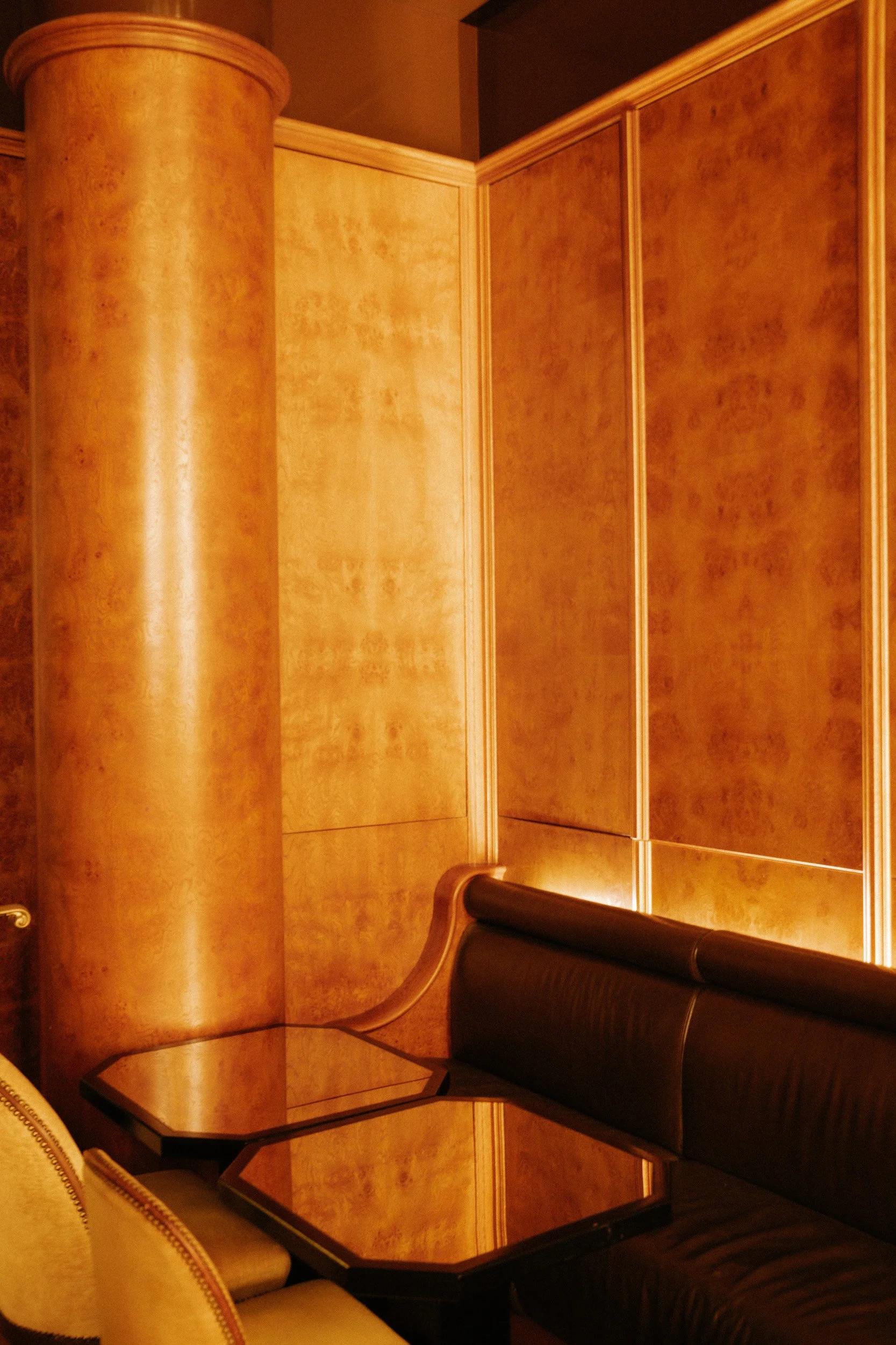 A corner of a wooden room with a built-in wooden cabinet or bar, a black leather bench, and a beige armchair at a wooden table.