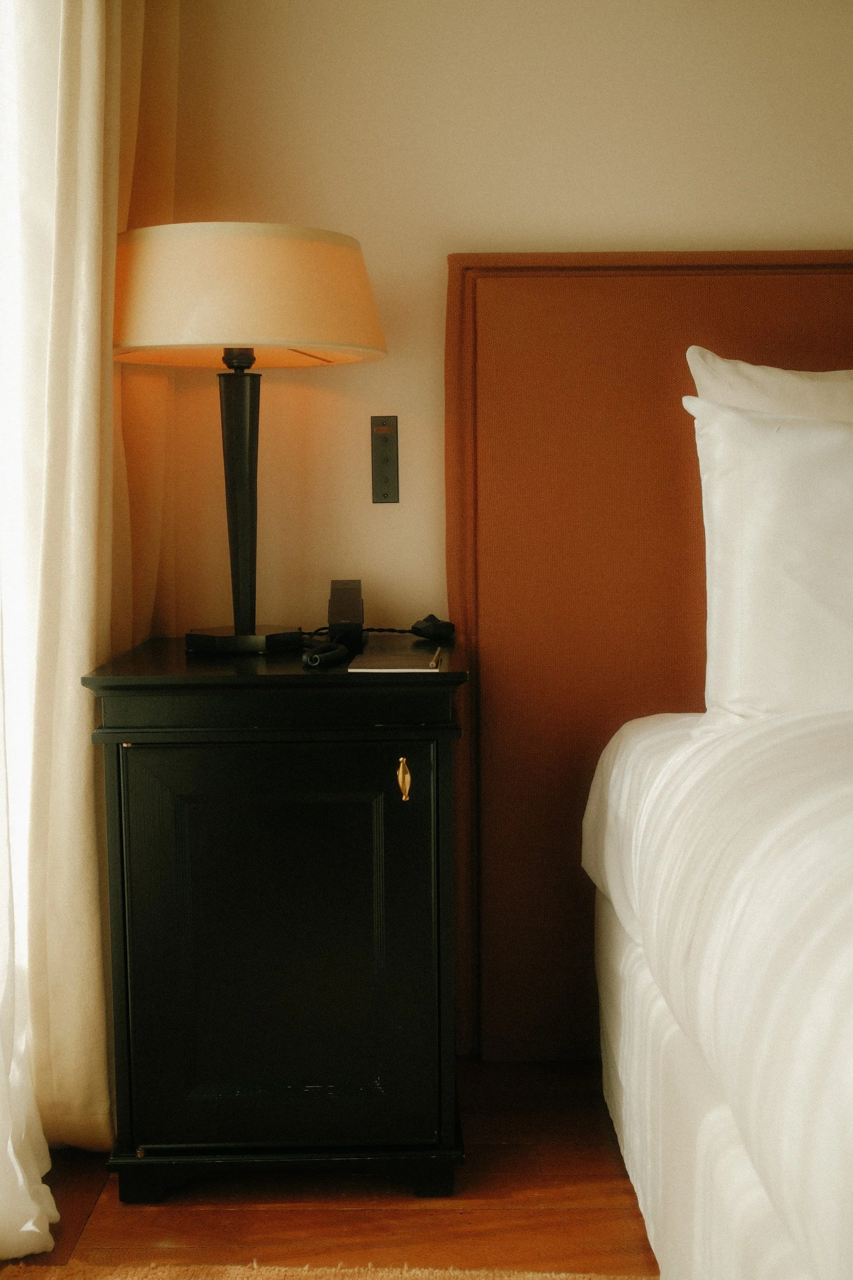 View of a hotel room corner showing a bed with white linens, a wooden headboard, a black nightstand with a lamp, a remote control, and a wall outlet, with a beige curtain.