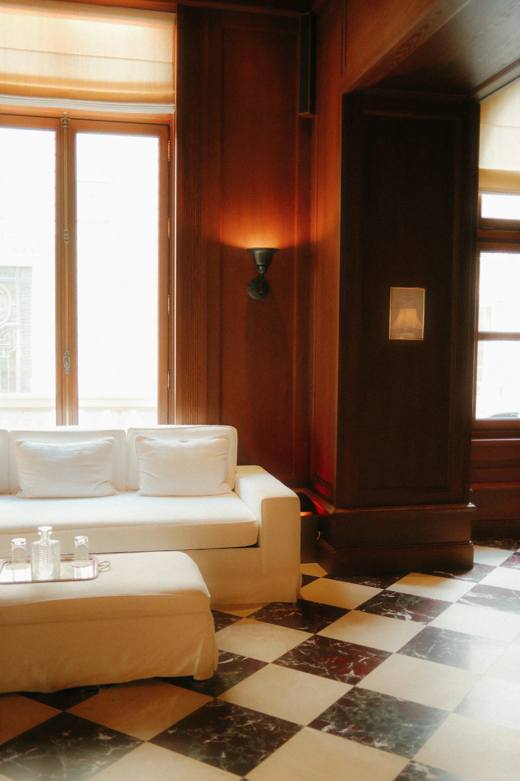 Interior of a room with a white sofa and a marble checkered floor, large windows with natural light, and wooden paneling on the walls.