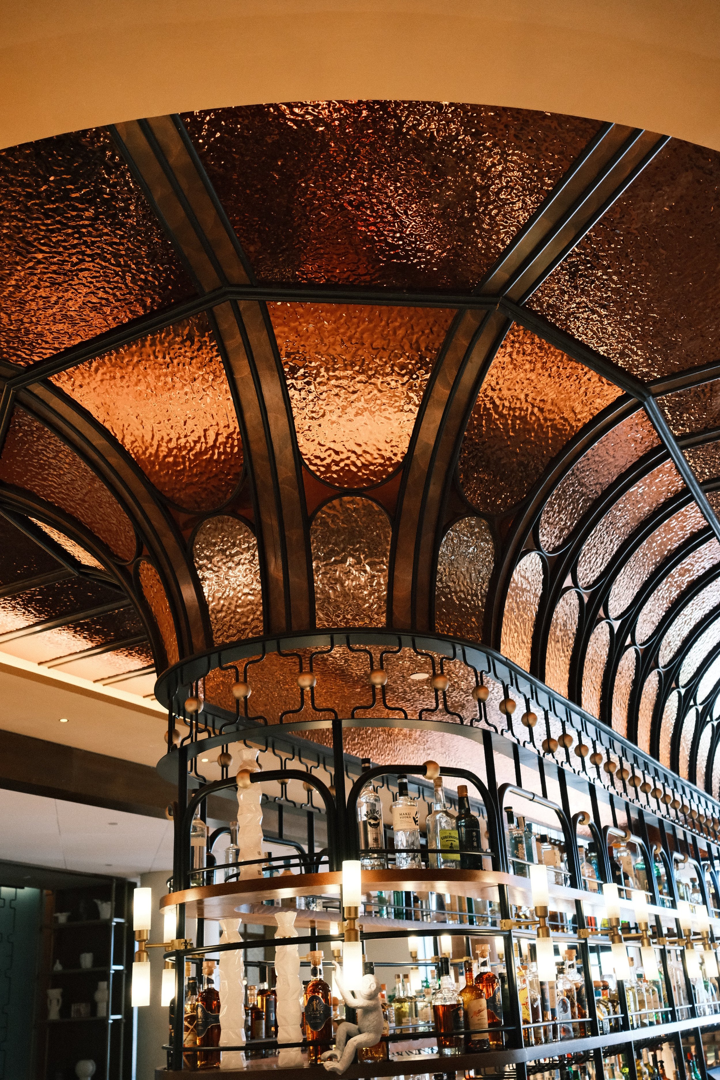 A rooftop bar with a curved, semi-transparent, textured orange canopy structure and a bar filled with various bottles of alcohol, illuminated by modern pendant lights.