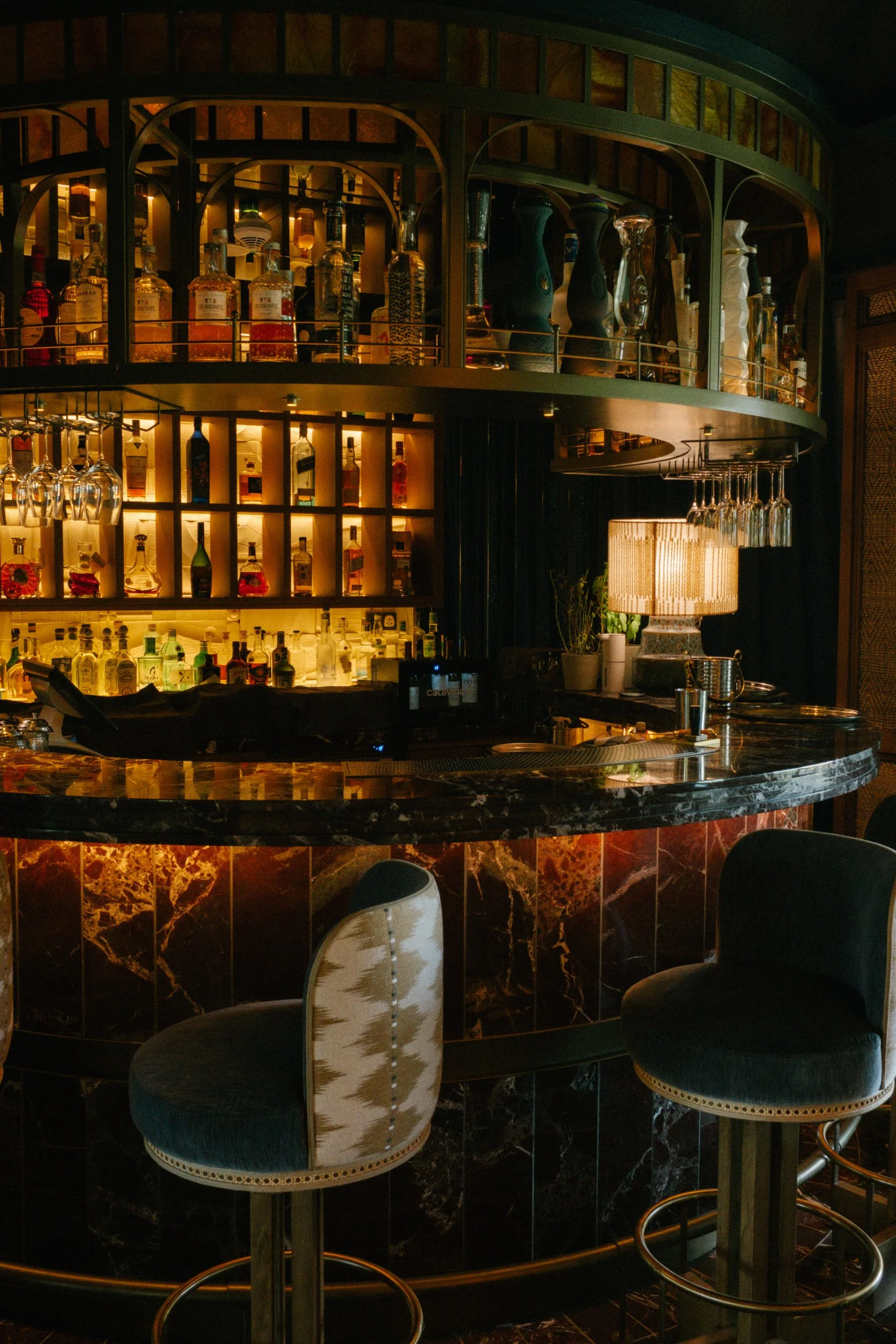 A bar area with a curved marble countertop, black and brown marble tiles, and a backdrop of illuminated bottles of various liquors on shelves, with bar stools and chairs around.