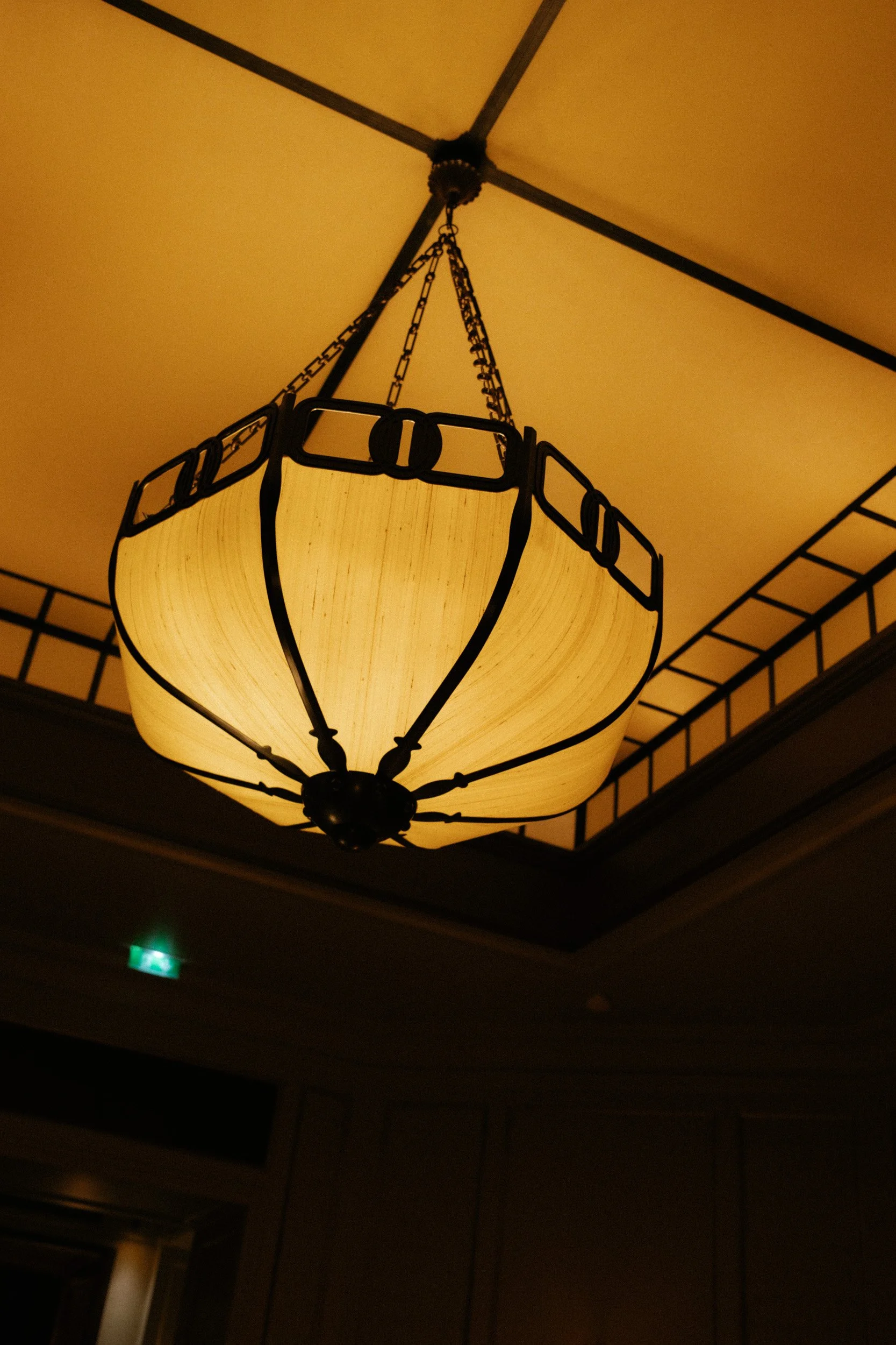 A hanging ceiling light with a yellowish lampshade, chains, and black metal accents. The ceiling is painted a yellow color, and there is an emergency exit sign illuminated green in the background.