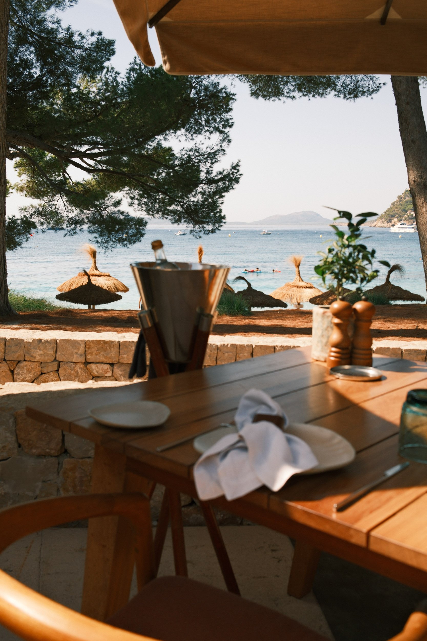 View of a beach with straw umbrellas and boats on the water, seen from an outdoor dining area with a wooden table set with plates and utensils.