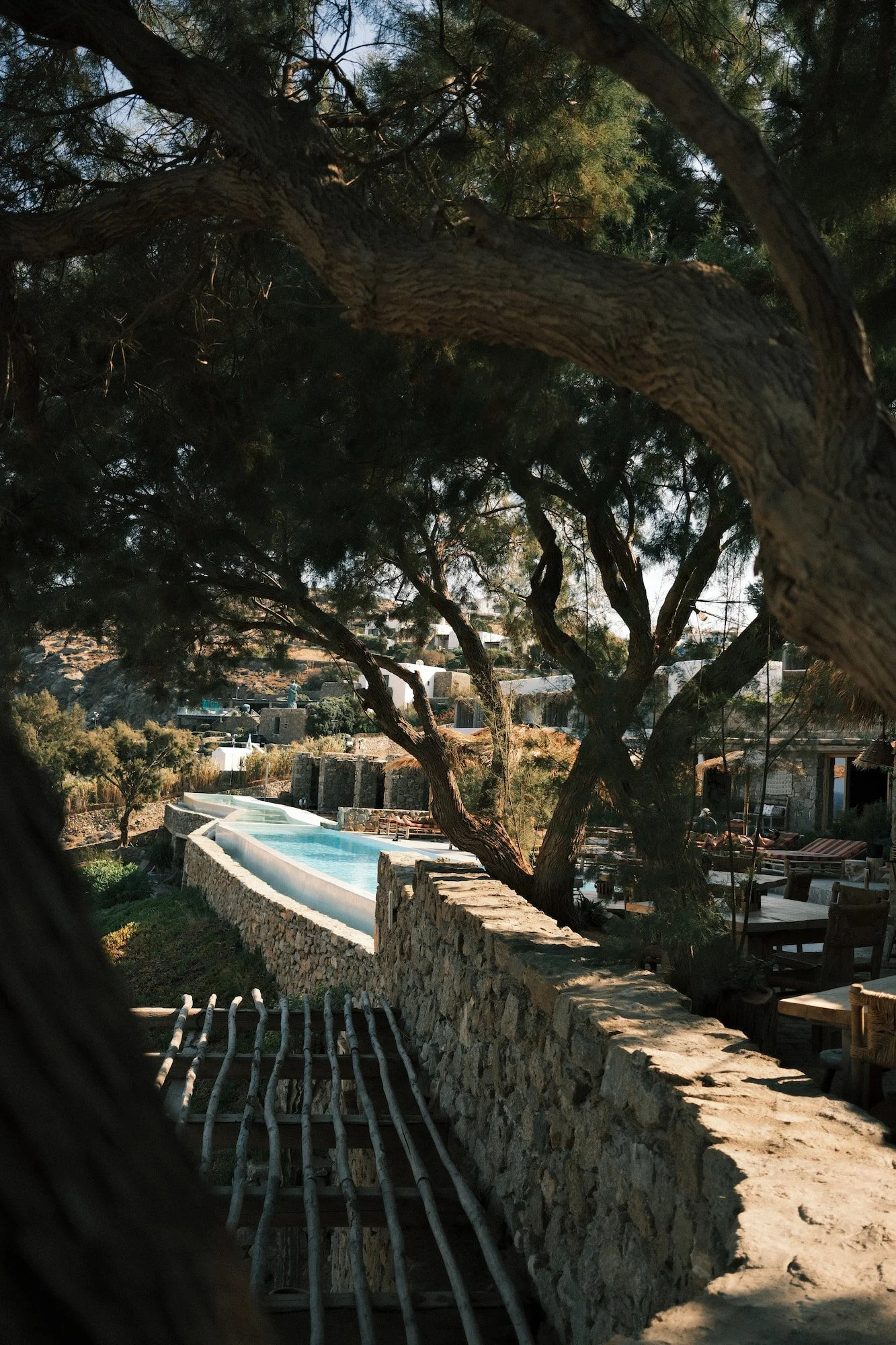 View of a backyard with a stone wall, a rooftop pool, trees, and a hillside with houses in the background, seen through a window or fence with dark framing.