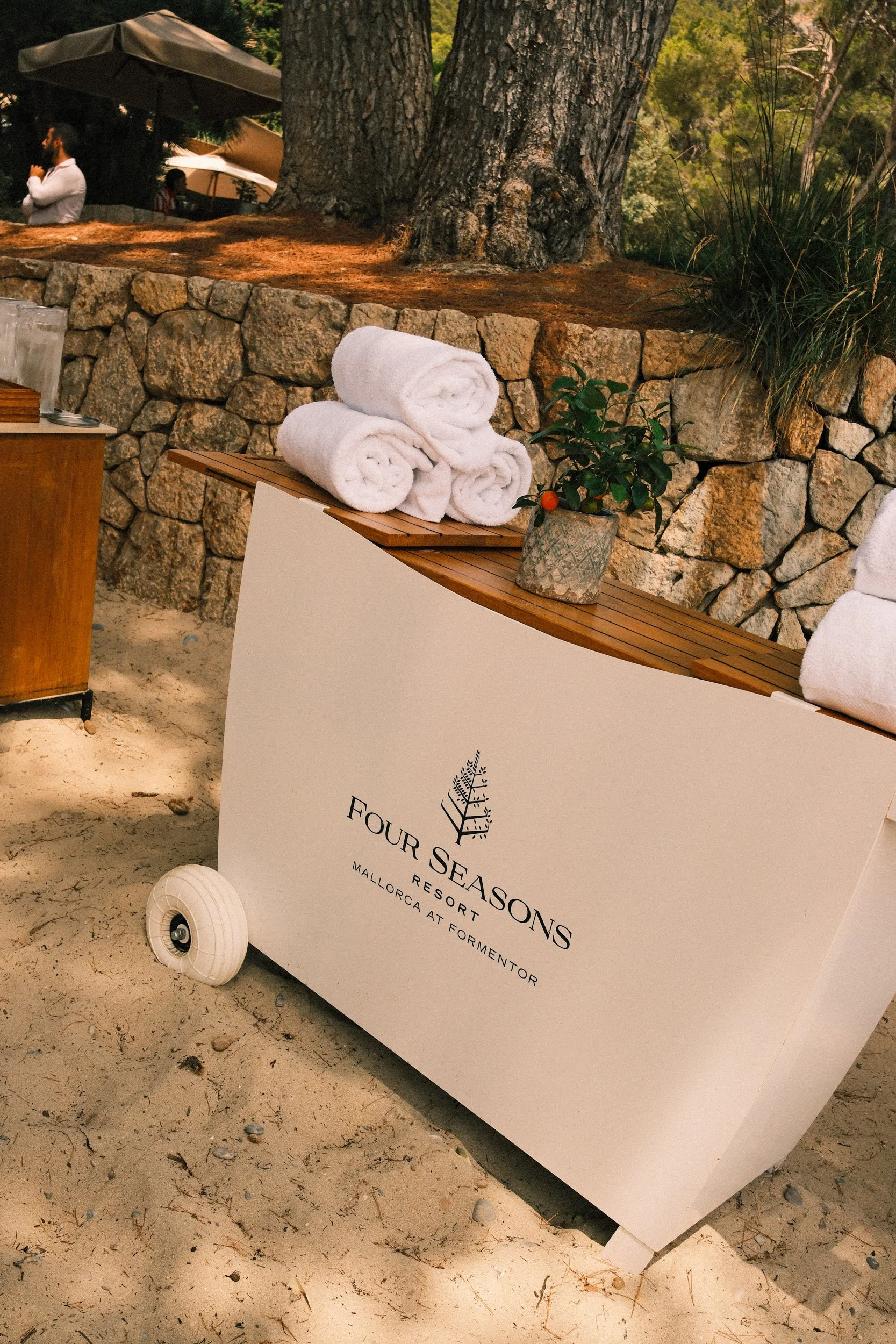 A white sign with black text that reads "Four Seasons Resort Mallorca at Formentor" leaning against a white stand on sandy ground. There are four neatly rolled white towels on a wooden bench, a small potted plant with green leaves and a few orange be