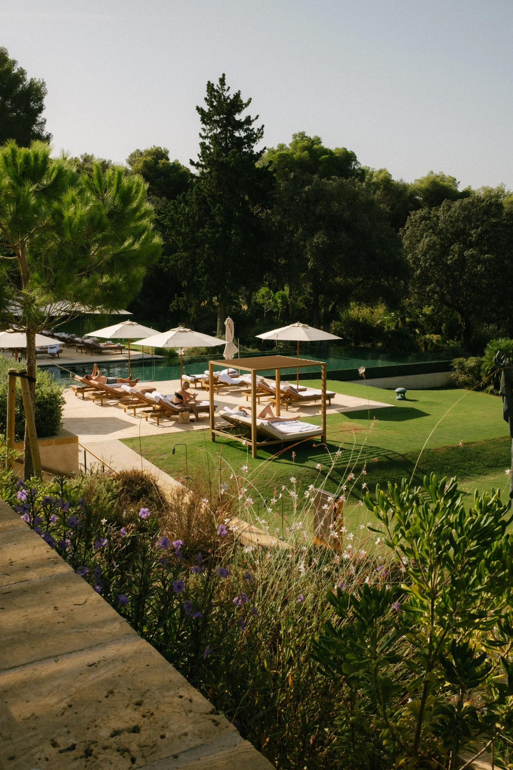 A poolside area with lounge chairs, white umbrellas, and a wooden cabana, surrounded by trees and greenery.