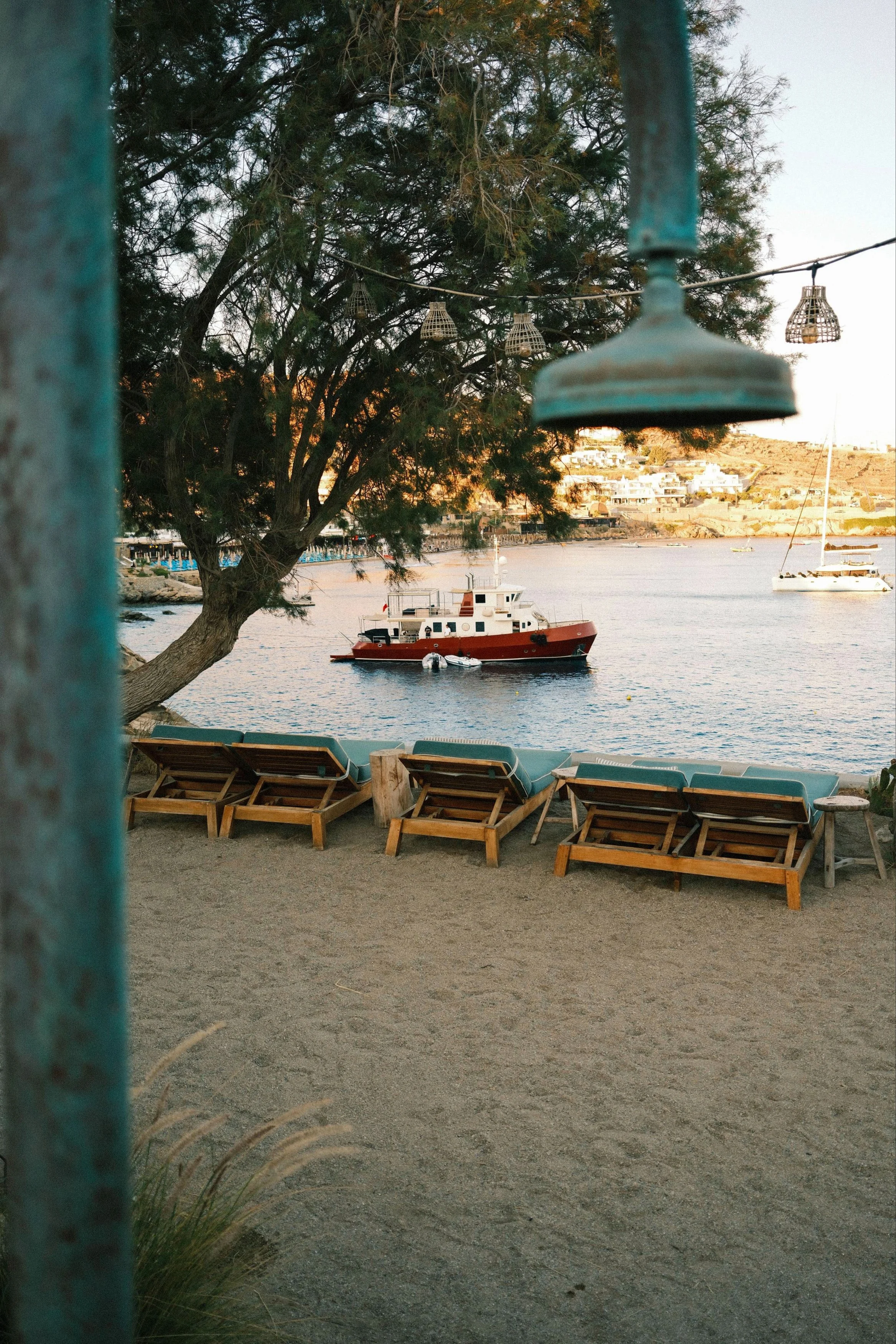 Beach scene with lounge chairs on sand facing the water, a dock with boats, a large tree with hanging lanterns, and a hill with houses in the background during sunset.