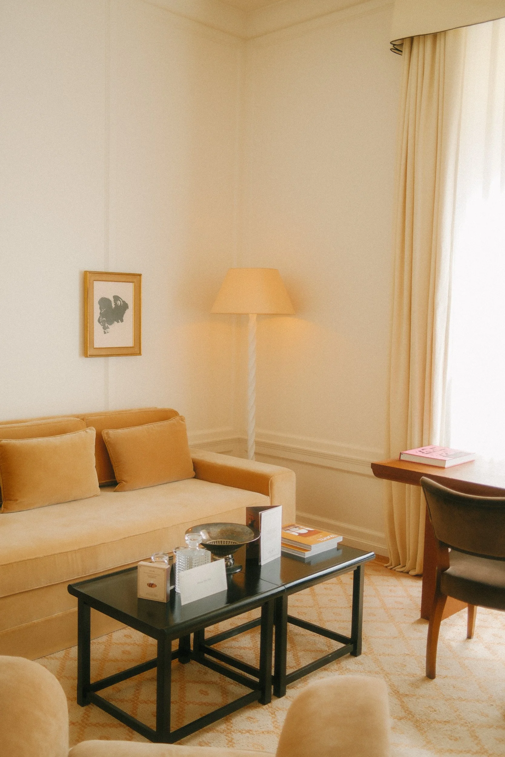 A cozy living room with a beige sofa and matching pillows, a black coffee table with books and decorative items, a lamp in the corner, cream-colored curtains, and a framed artwork on the wall.