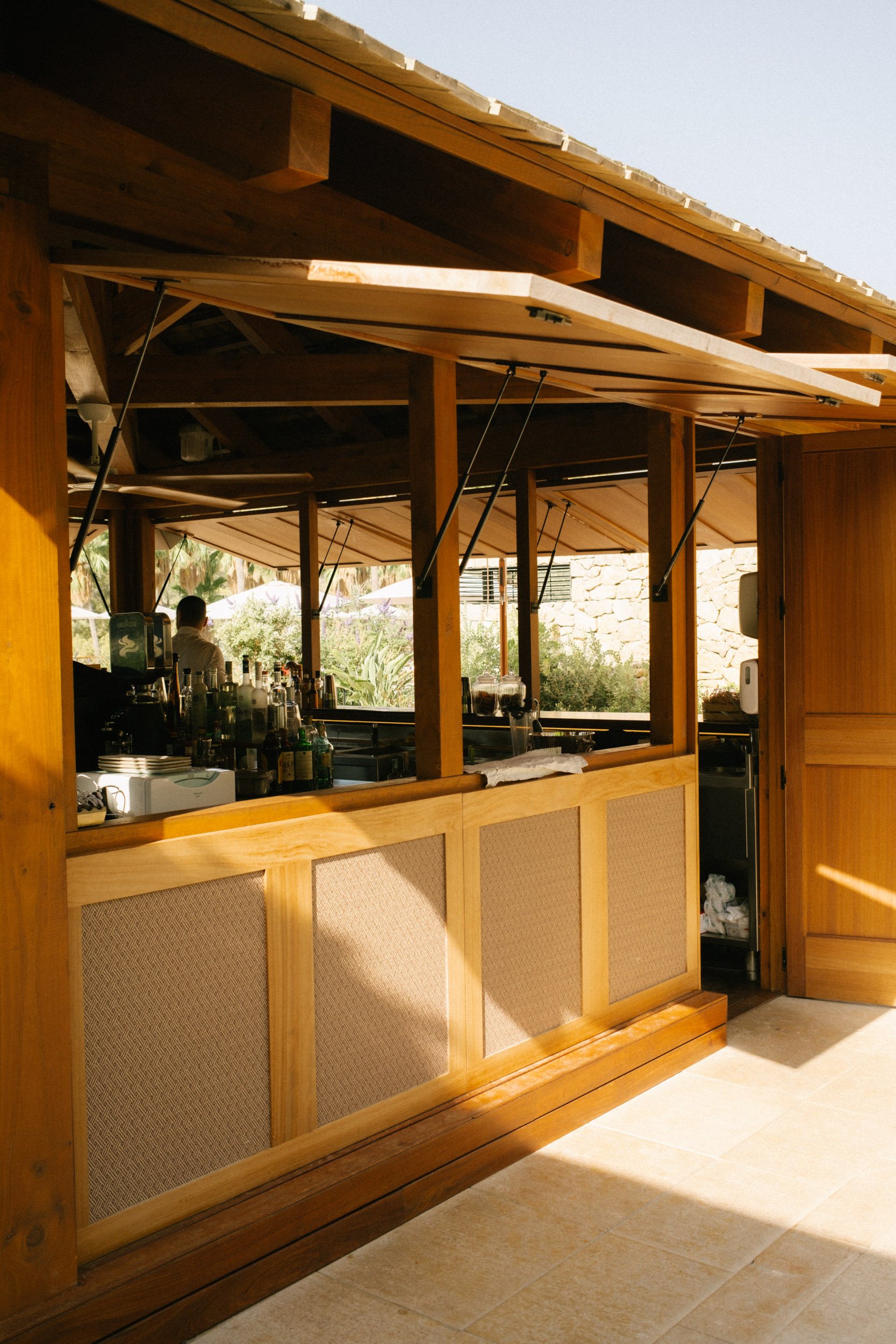 Wooden bar counter with beige panels and natural finish, situated inside a room with a tiled floor. Background shows lockers, a man, and tropical plants outside.