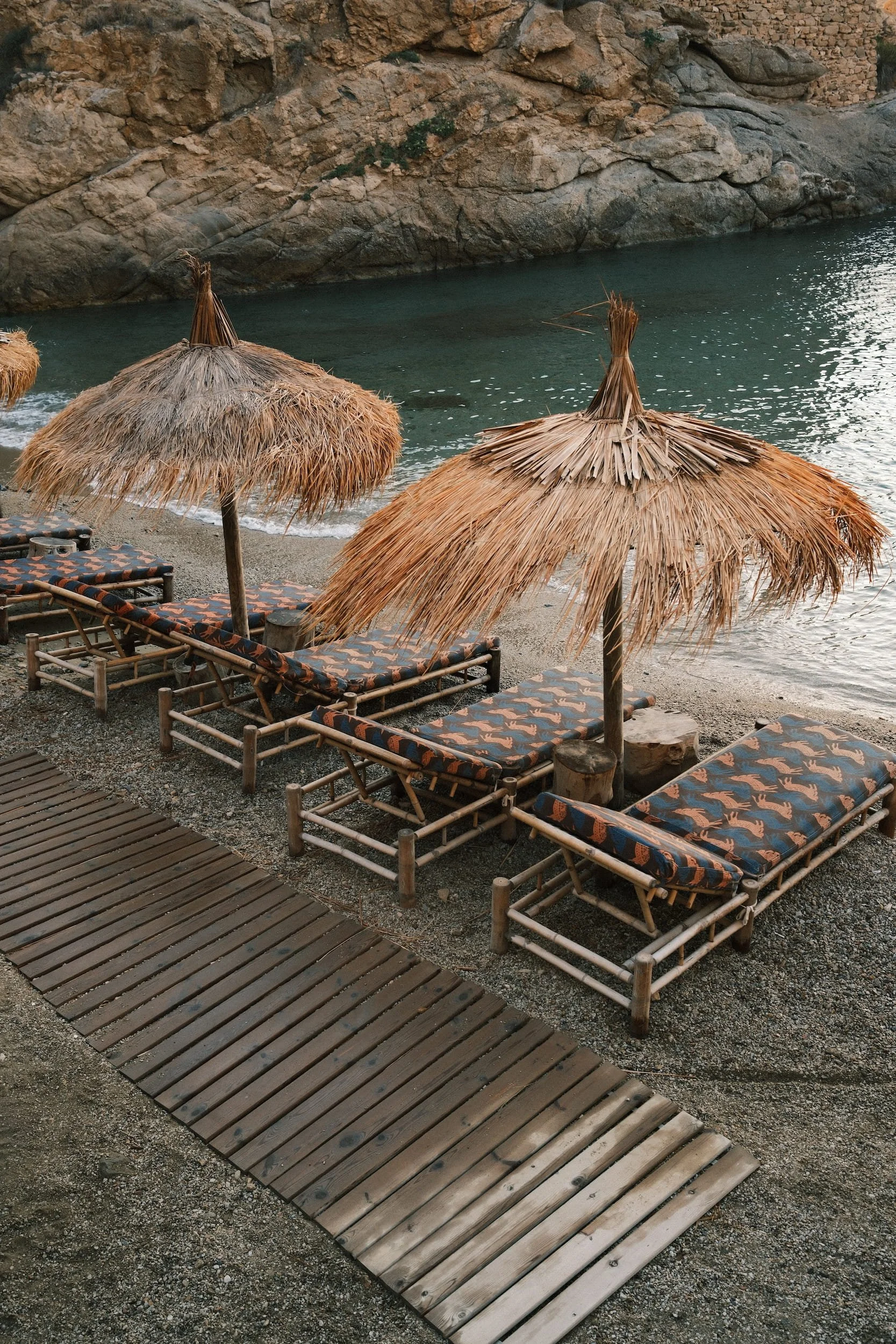 Beach scene with three straw umbrellas over patterned lounge chairs along the shoreline, with rocky cliffs in the background and calm water.