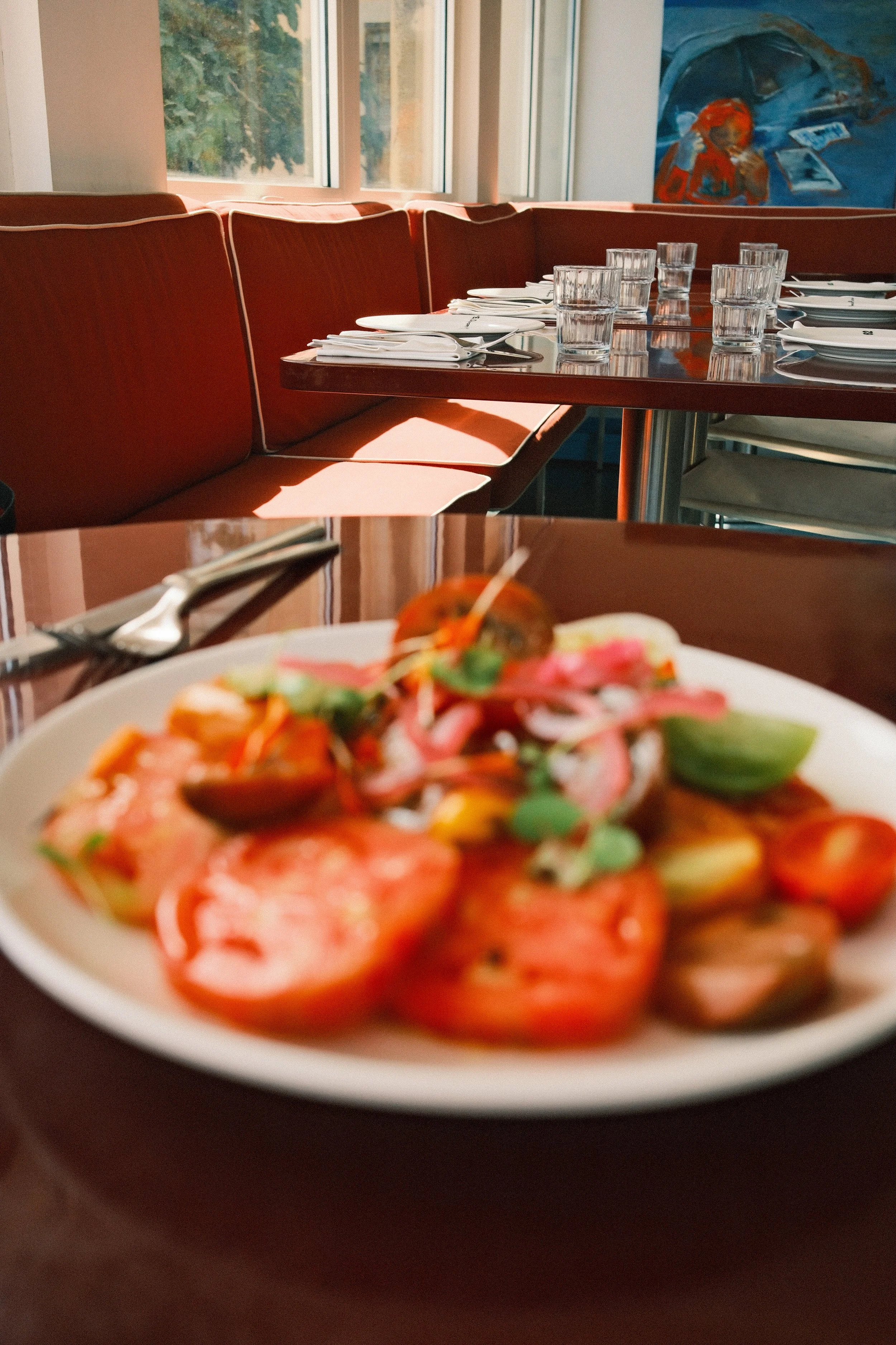A plate of salad with fresh tomatoes, greens, and other vegetables, placed on a dark table in the restaurant. In the background, there are table settings with glasses and silverware, and a painting on the wall.