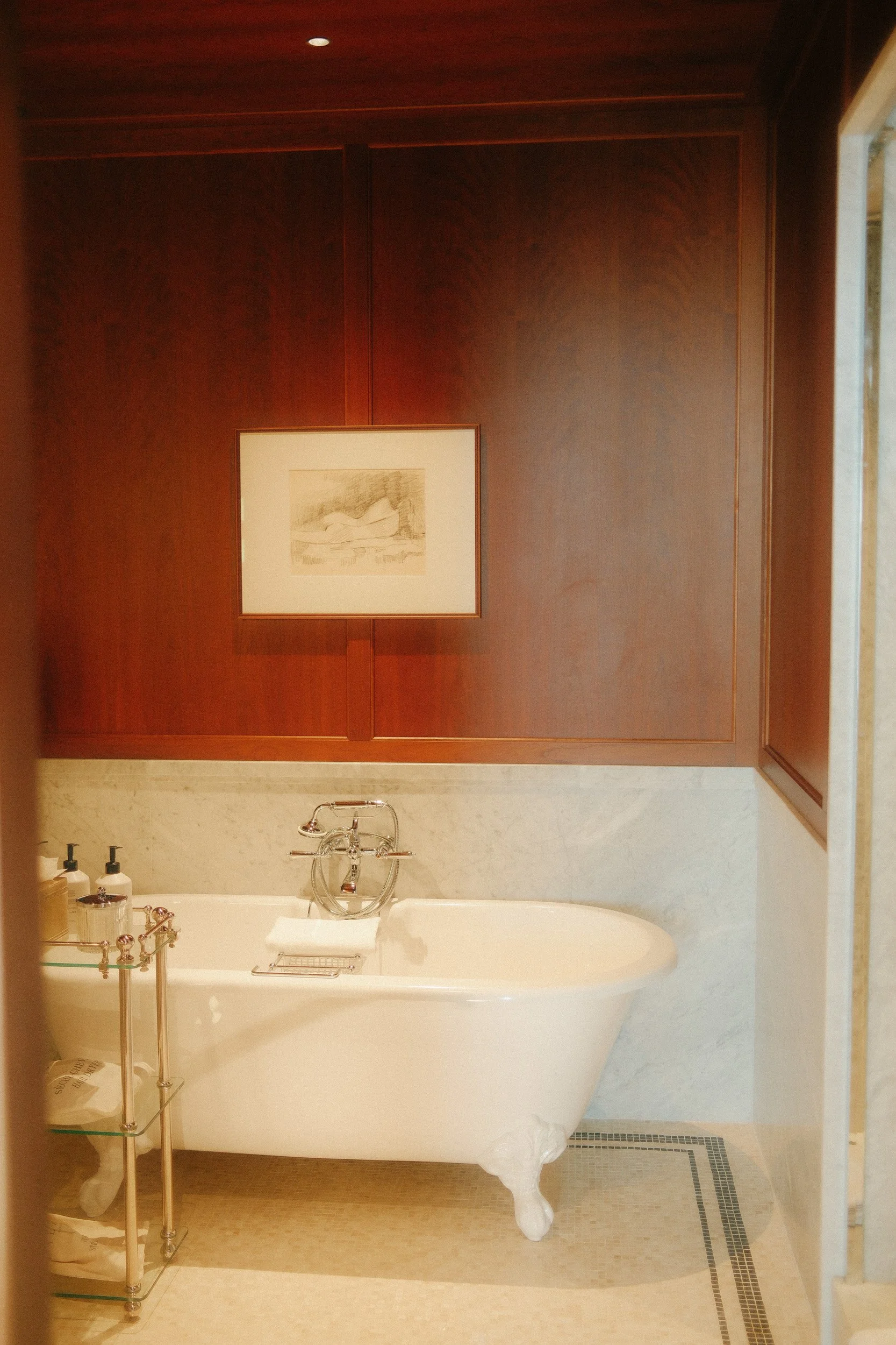 A bathroom with a white clawfoot bathtub, a wooden wall with a framed sketch, a small shelf with toiletries, and a window to the right.