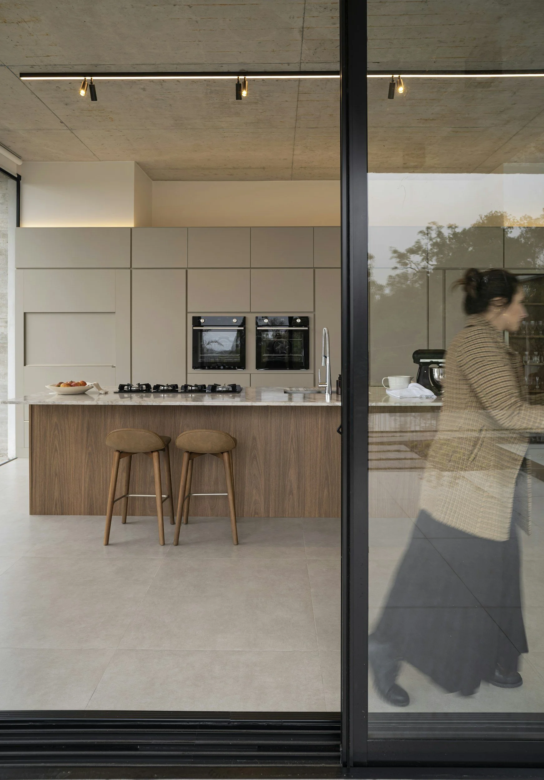 Modern kitchen with a beige island, two brown stools, white cabinetry, and built-in ovens. A woman wearing black shoes and a beige checked coat walks past a glass wall that shows trees outside.