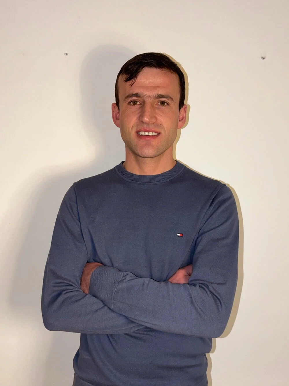 A man with short dark hair, wearing a navy blue long-sleeve shirt with a small logo on the chest, stands with his arms crossed in front of a plain white wall.