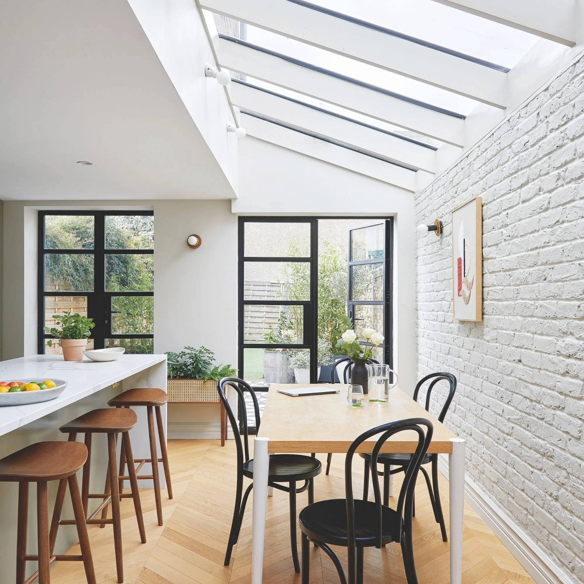 Interior of a bright dining area with large skylight windows, white brick wall, black framed windows, and a wooden floor. There's a light wooden dining table with black chairs, a pot with white roses, and a bowl of fruit.