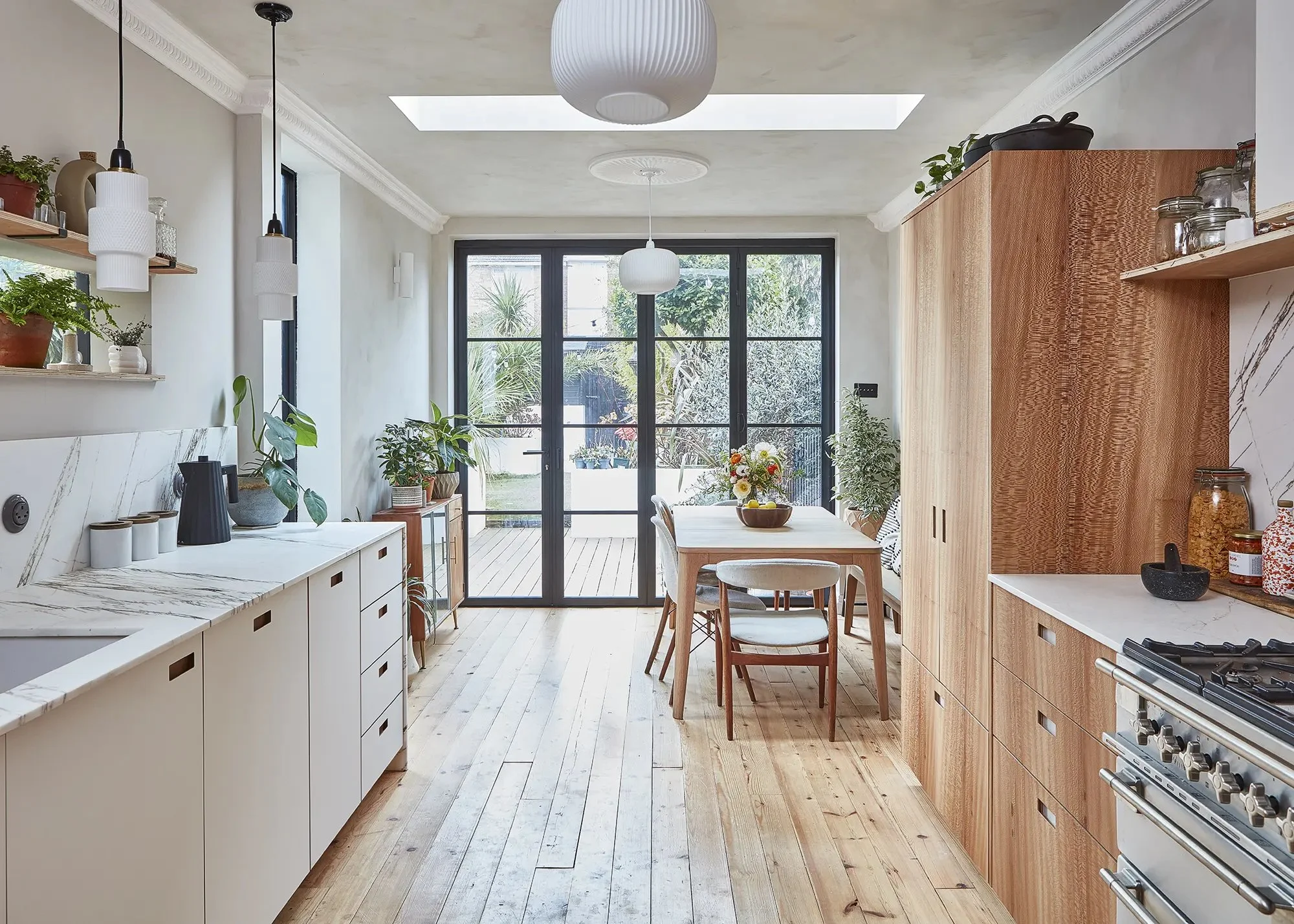 Modern kitchen with wooden and white cabinets, plants, hanging pendant lights, and large glass doors leading to an outdoor area with trees and plants.