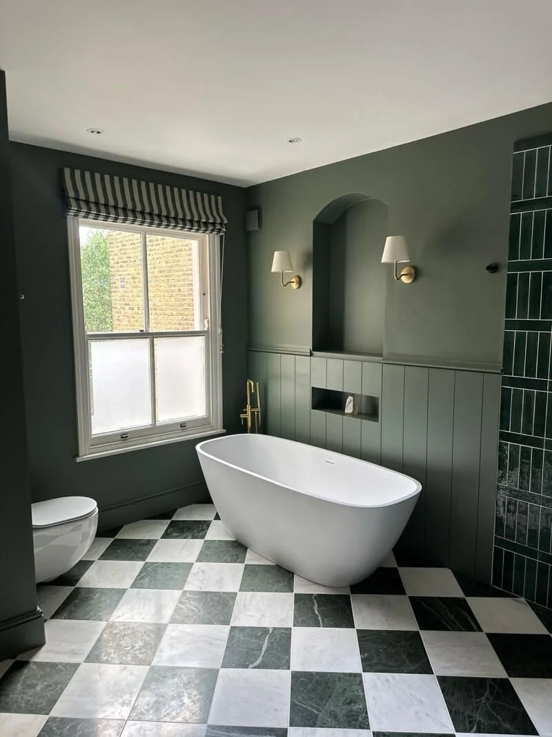 Bathroom with black and white checkered tile floor, green walls, a white freestanding bathtub, a toilet, a window with a striped valance, and gold fixtures.