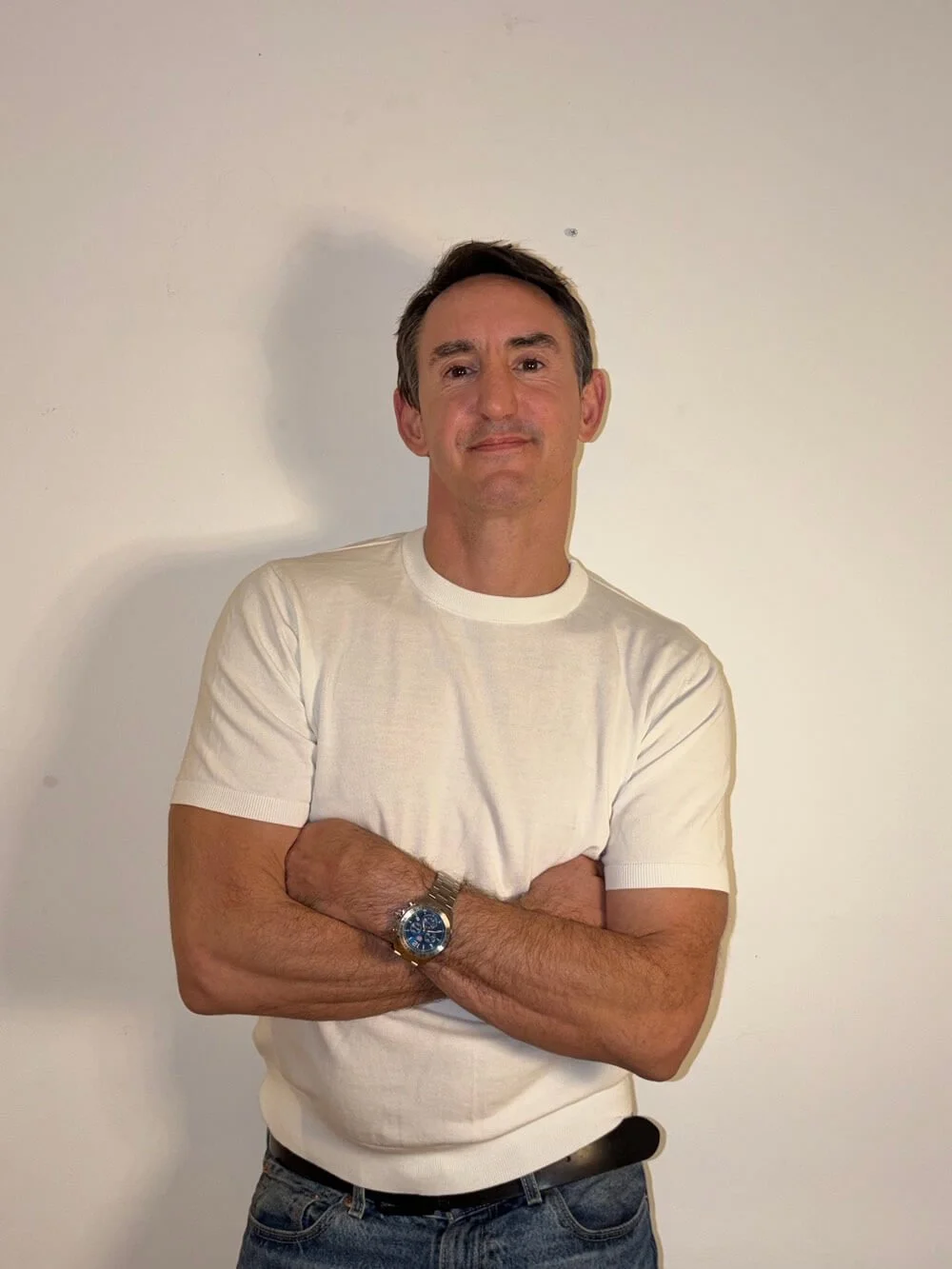 A man with dark hair wearing a white short-sleeve shirt and jeans, crossing his arms and standing against a plain white wall.