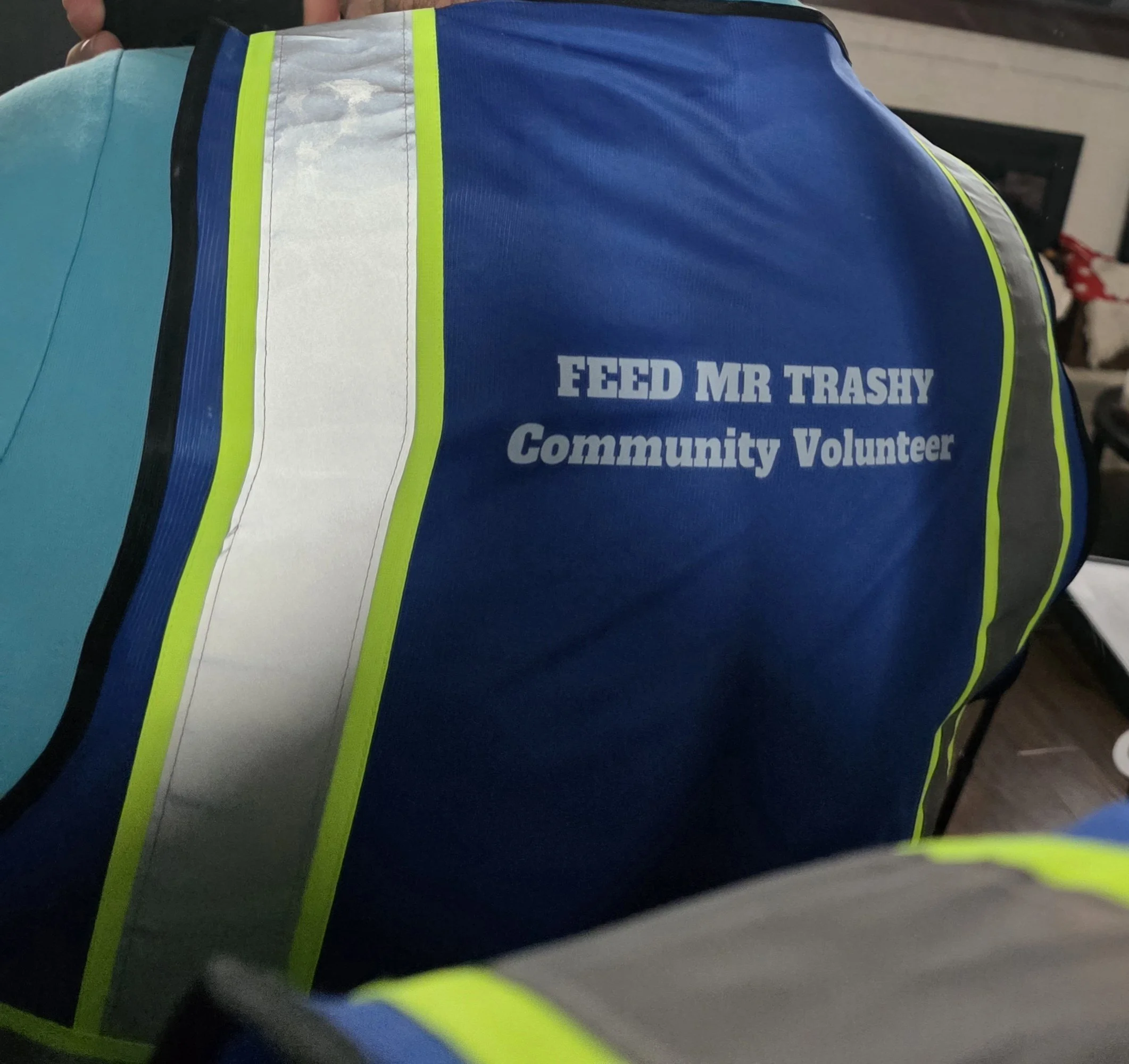 Close-up of a reflective safety vest with the text 'FEED MR TRASHY' and 'Community Volunteer' on it, worn by a person.