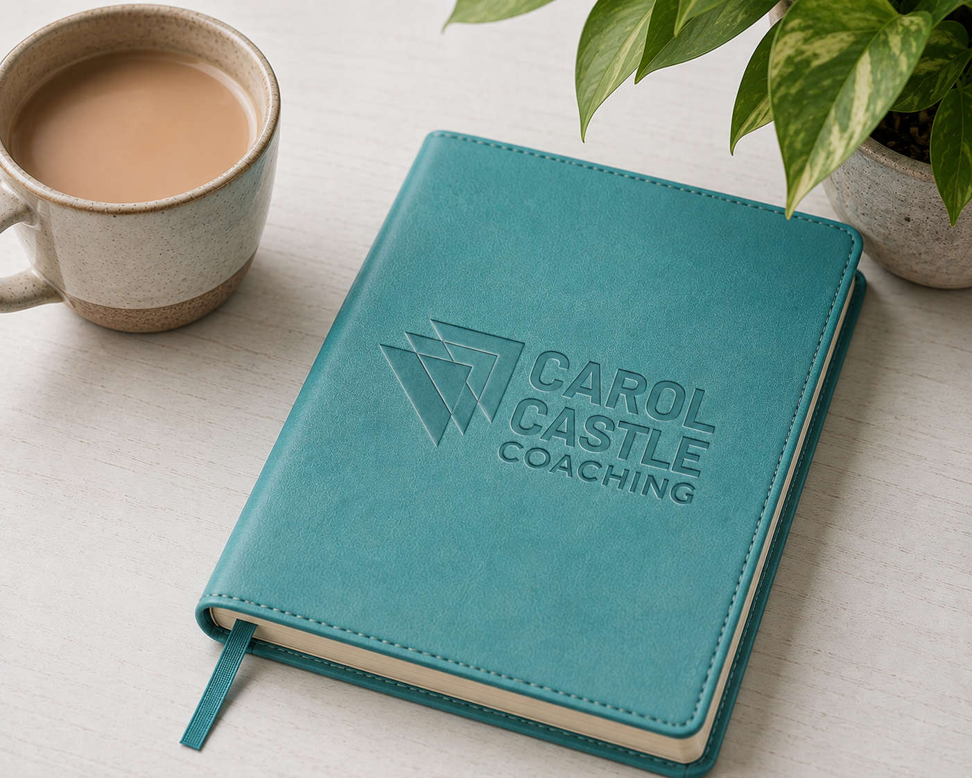 A teal notebook with the text 'Carol Castle Coaching' and a logo on the cover, a cup of coffee or tea, and a potted plant on a white desk.