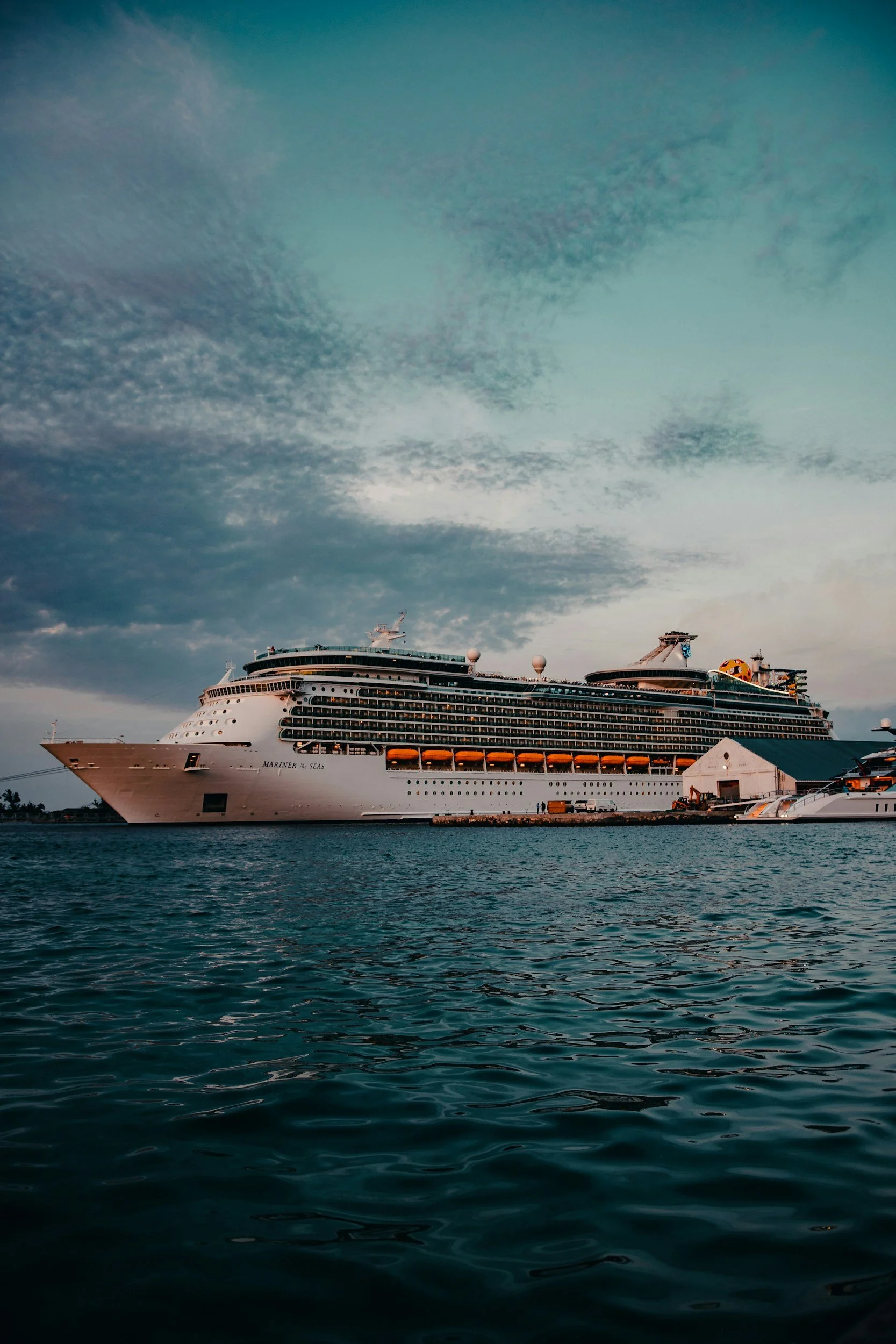 Large white cruise ship docked at a harbor with dark water in the foreground and a partly cloudy sky overhead.