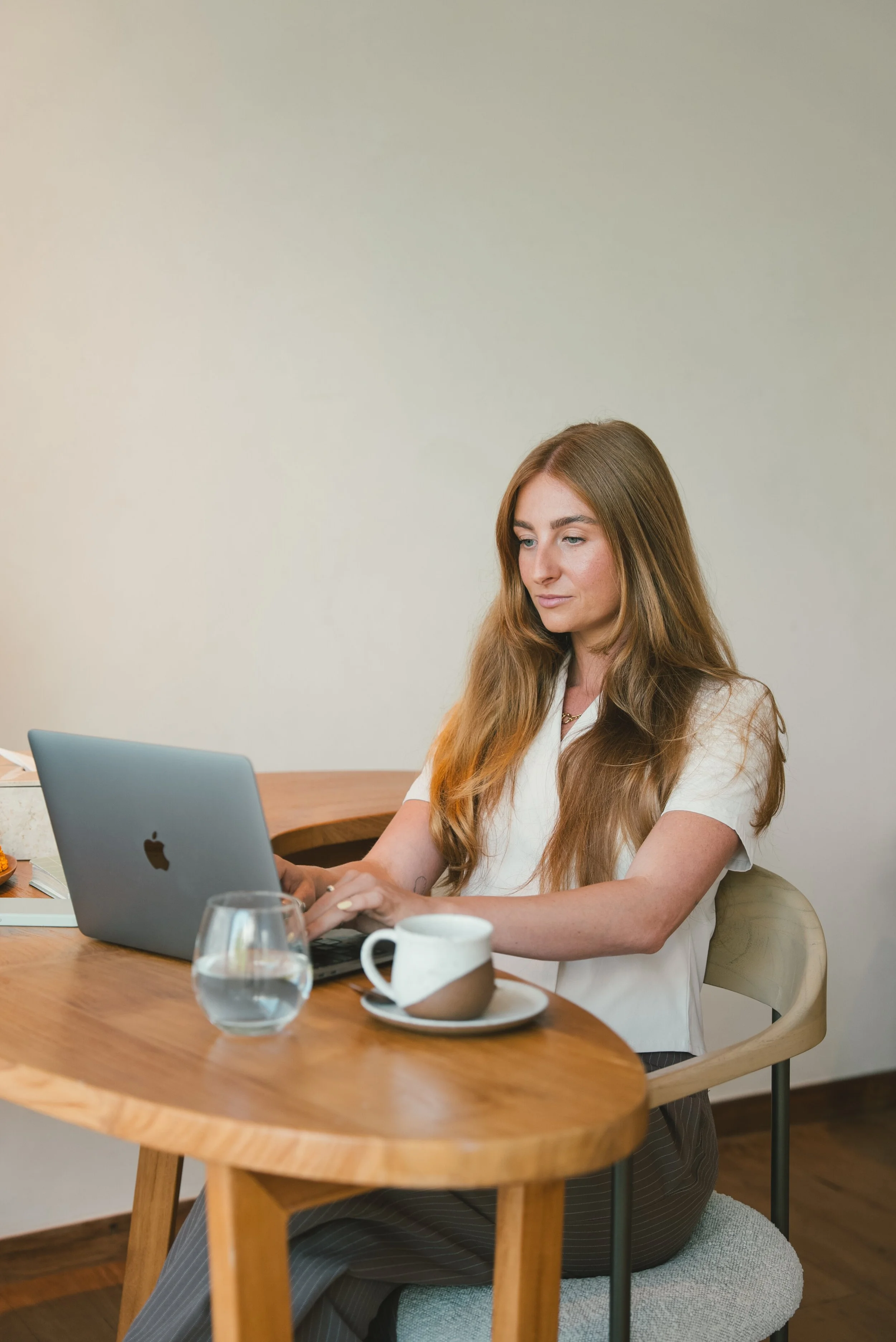 A woman with long reddish-brown hair working on a laptop at a wooden dining table in a modern, minimalistic room.