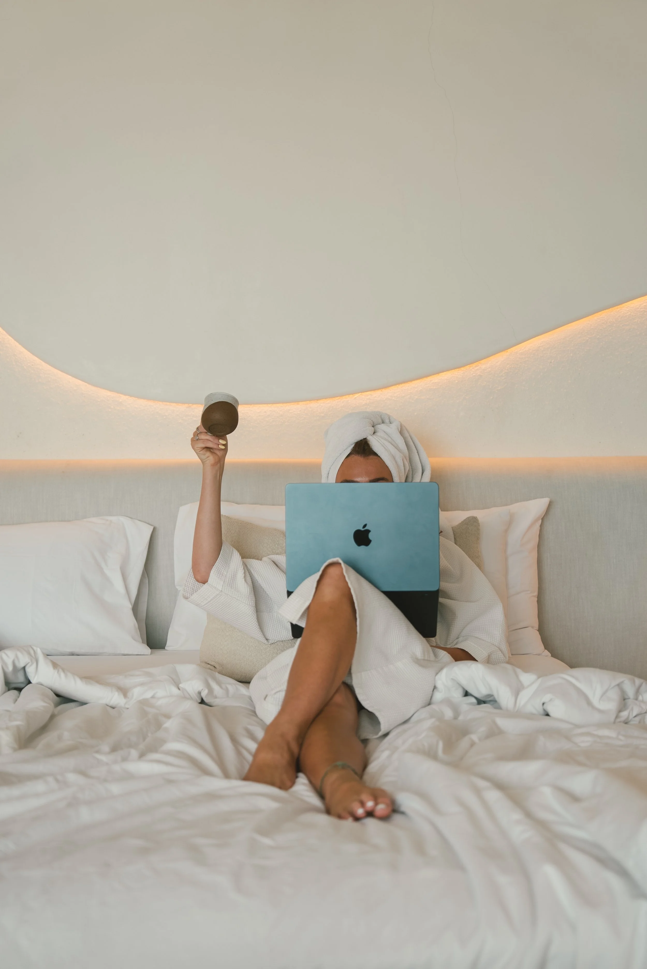 Woman in a white bathrobe with a towel wrapped around her head sitting on bed using a laptop, with a glass of coffee in hand, in a modern bedroom with beige curtains and illuminated ceiling design.
