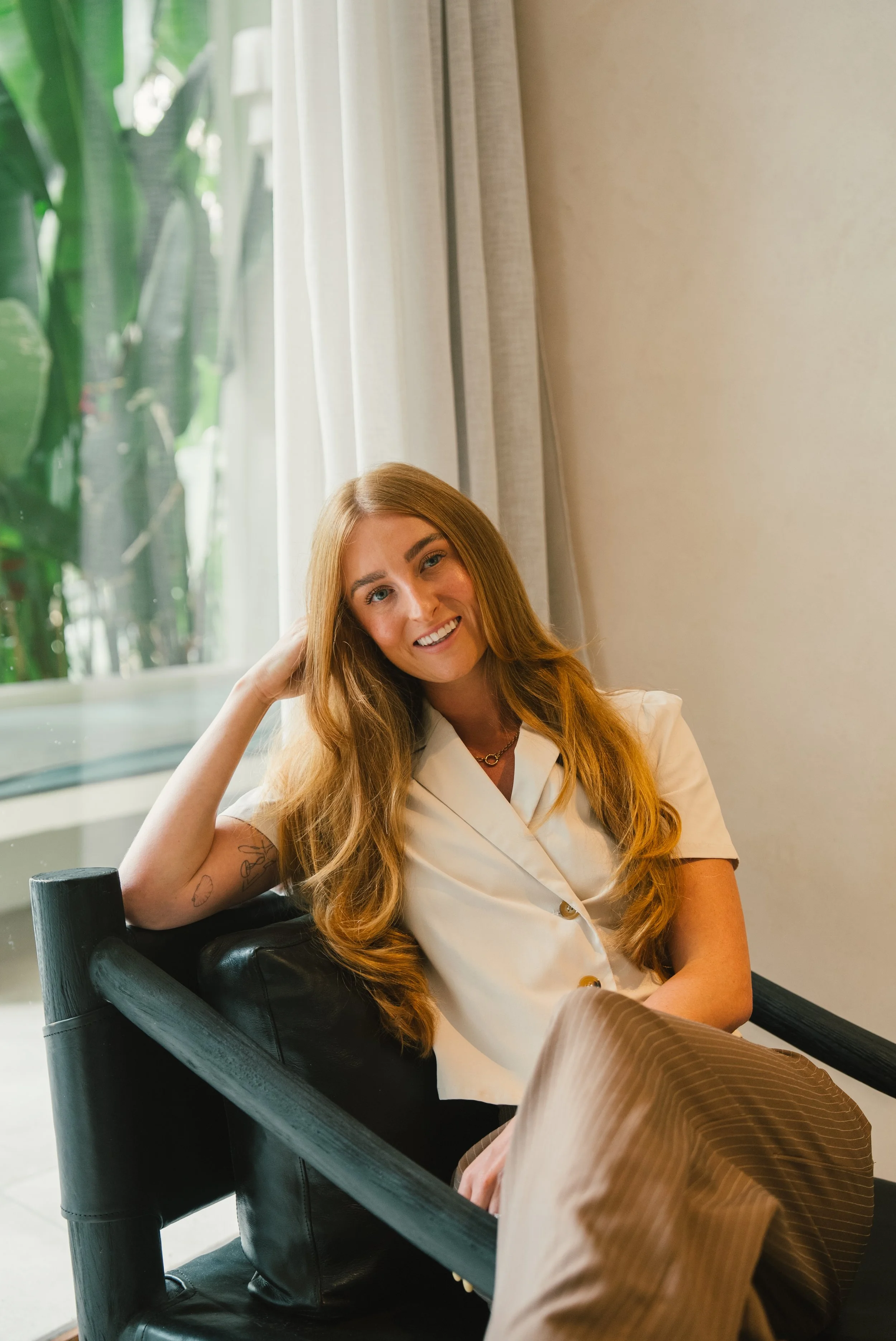 A woman with long red hair smiling and sitting in a black chair near a window with white curtains and green foliage outside.