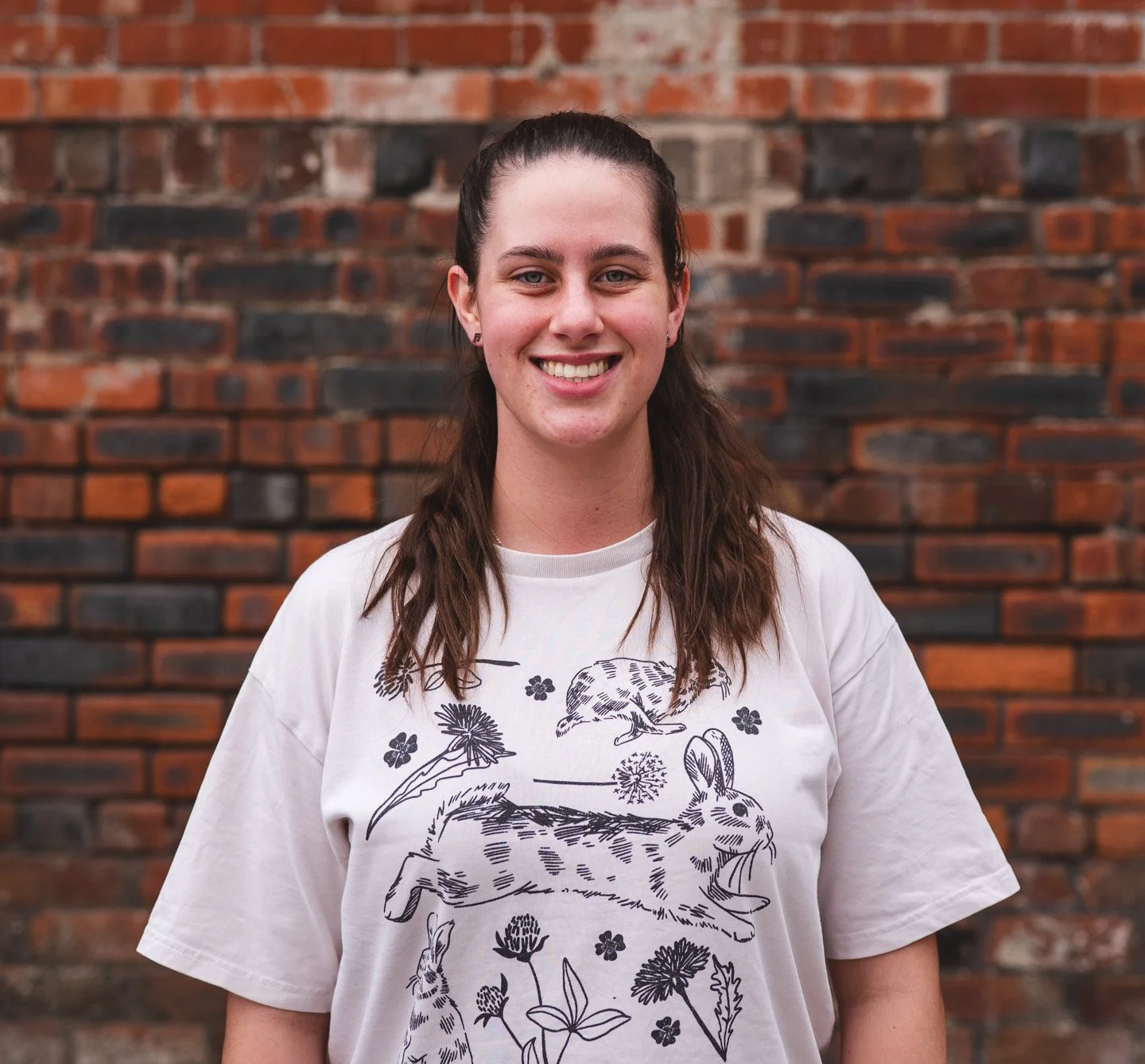 A young woman with brown hair tied back, smiling, wearing a white graphic T-shirt with rabbit and nature illustrations, standing in front of a brick wall.