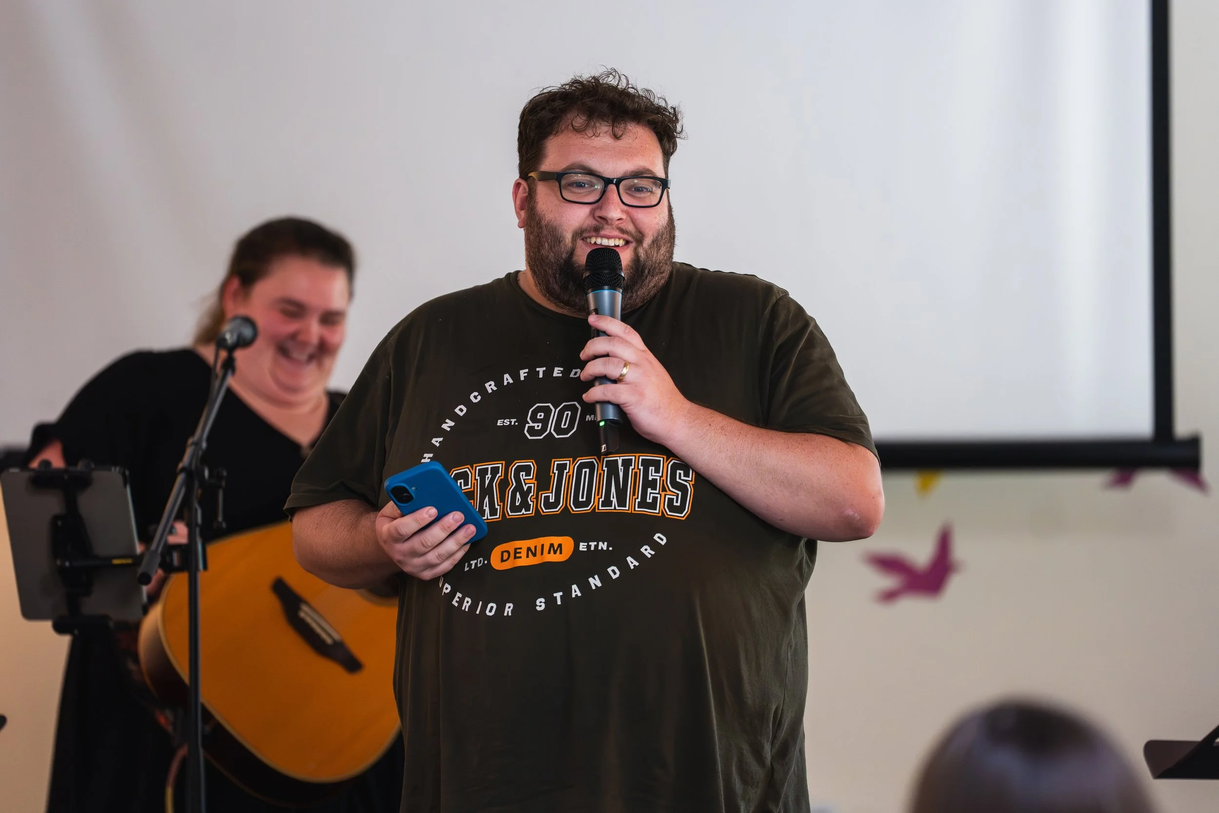 A man with glasses, beard, and short curly hair holding a microphone and smiling, while a woman behind him is playing a guitar and also smiling.
