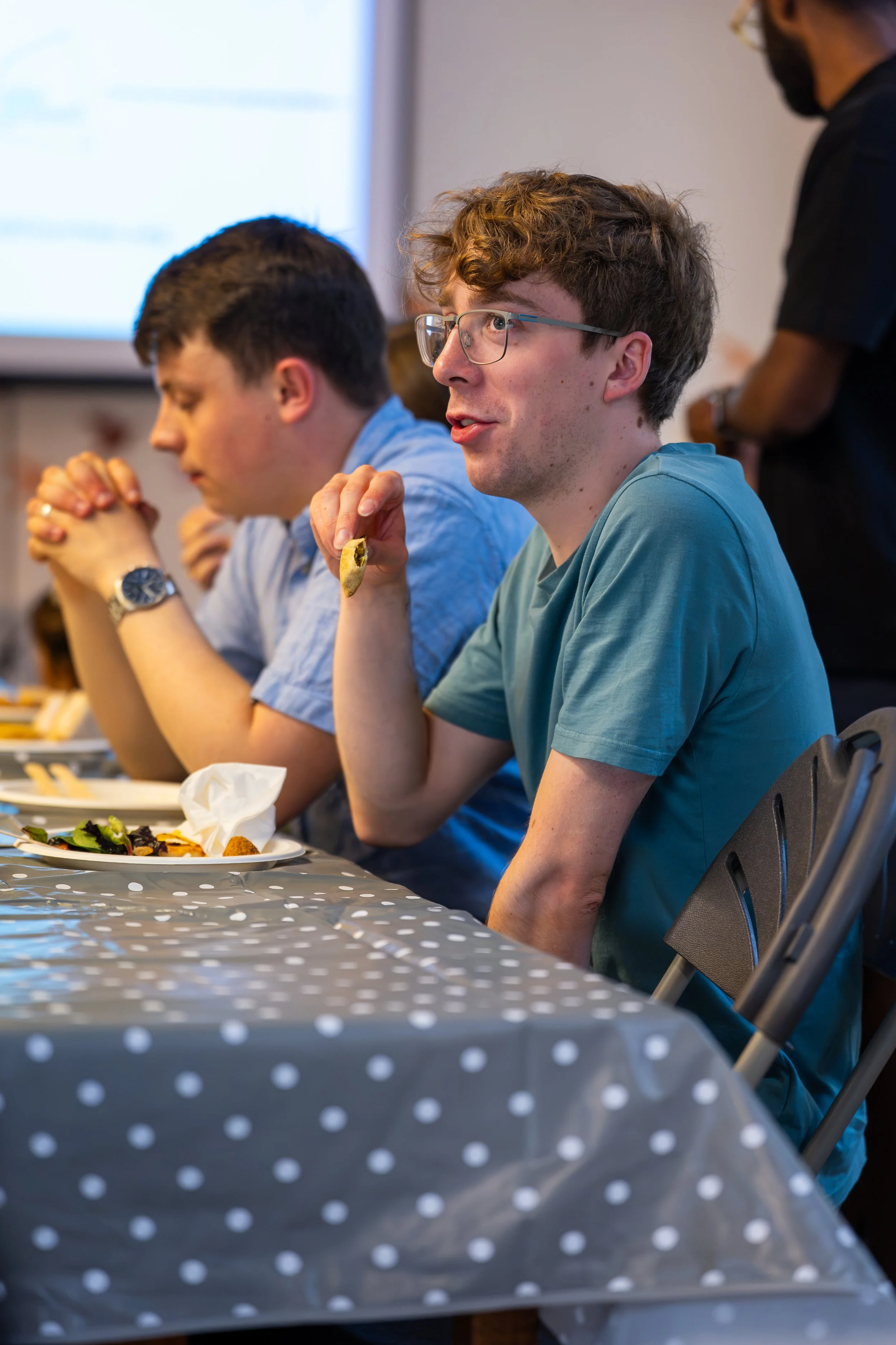 Young man with glasses eating a cookie at a table during a community event.