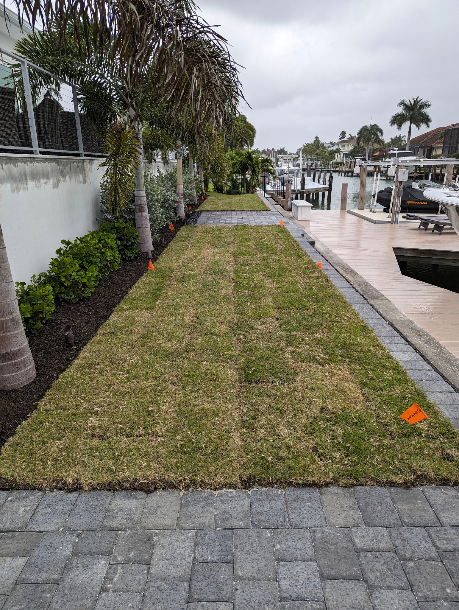 A landscaped yard with grass, palm trees, and shrubs on the left side, with a gravel and paver walkway. On the right, there is a dock with boats and a wooden deck, with houses and palm trees in the background. Overcast sky.