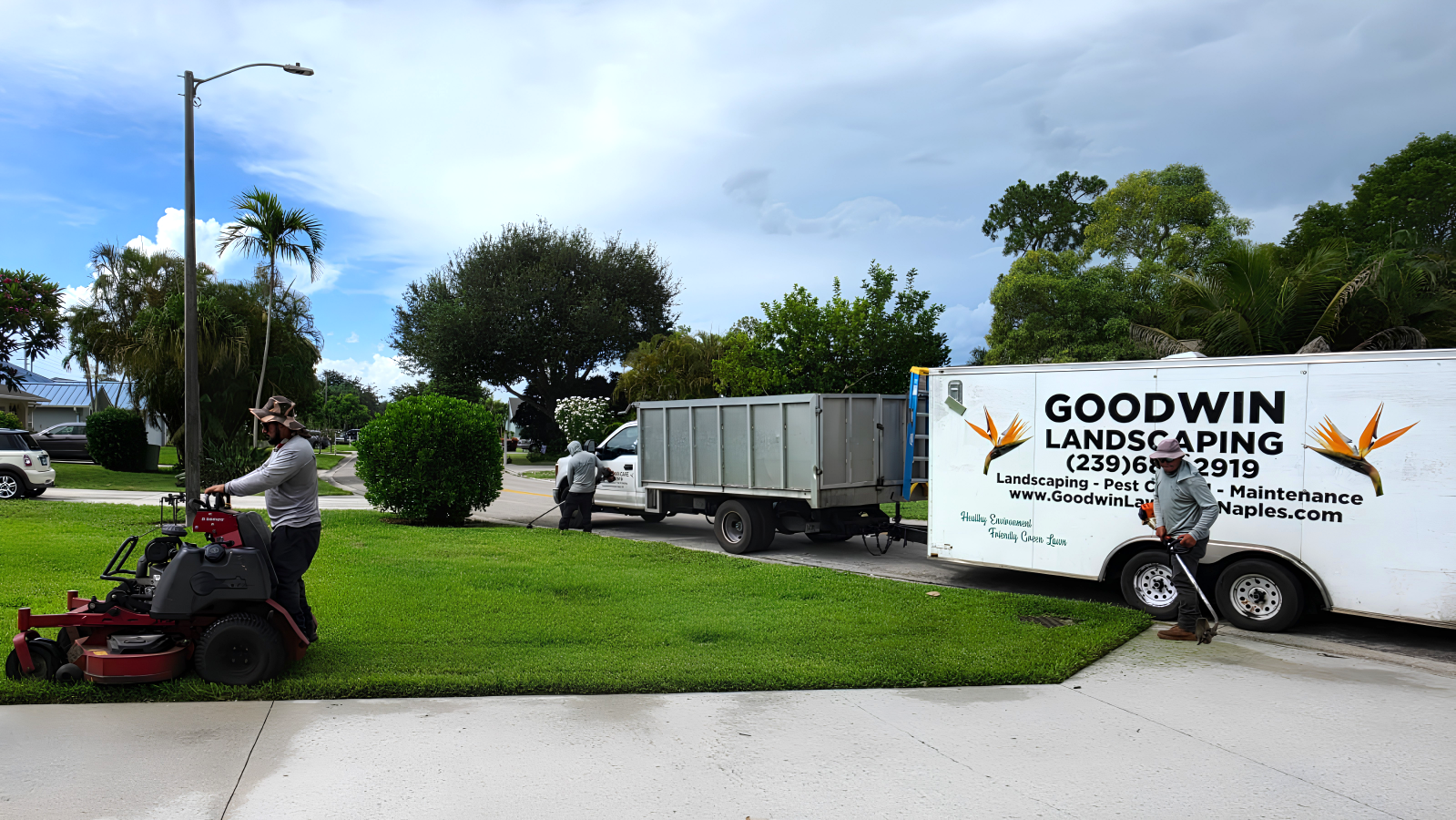 Two workers from Goodwin Landscaping trimming grass in a residential yard. One is using a riding mower and the other is using a handheld trimmer. There is a landscaping truck parked on the street behind them, with trees and houses in the background.
