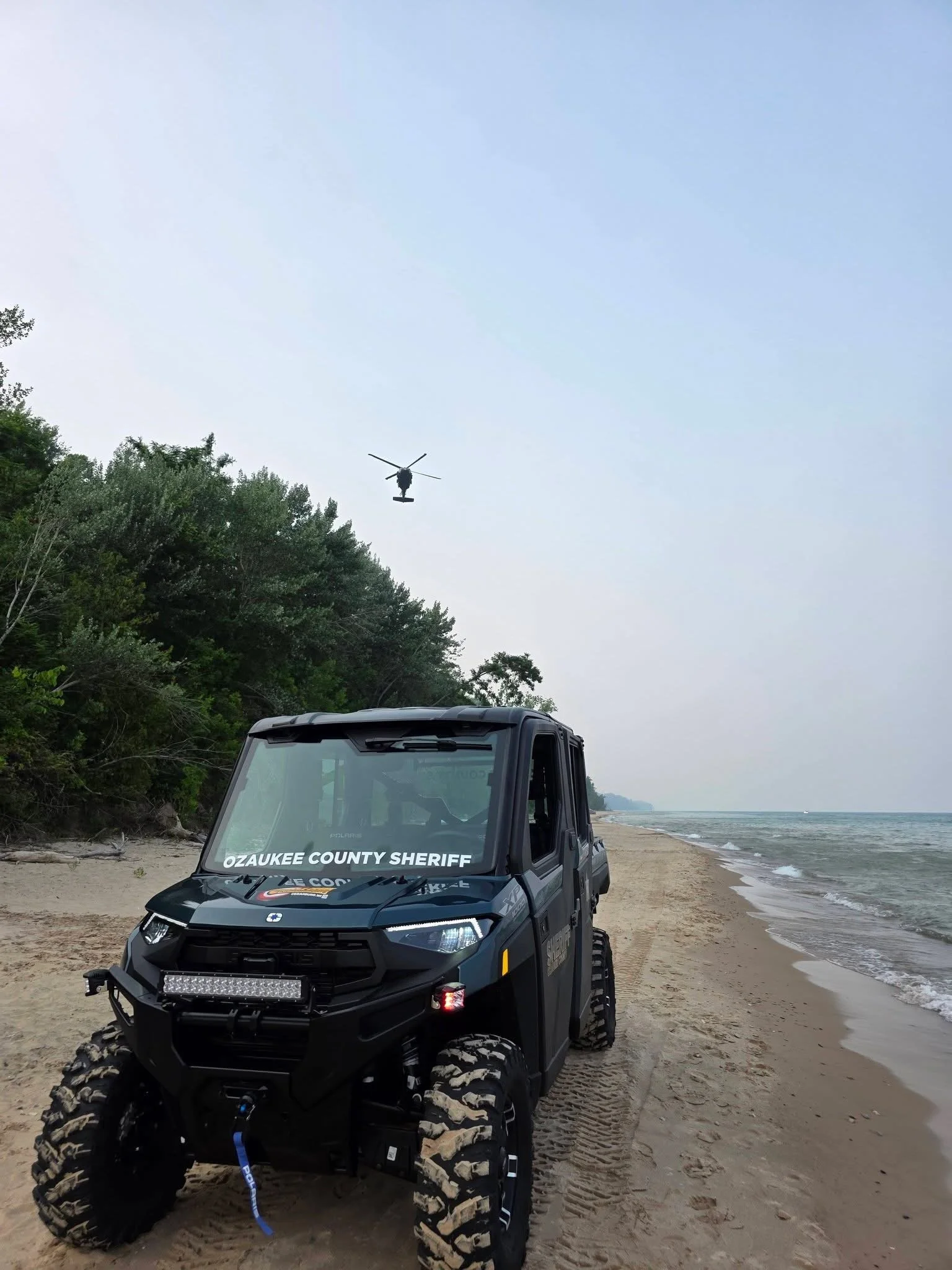 A black off-road utility vehicle with 'OZAKEE COUNTY SHERIFF' written on the windshield, parked on a sandy beach near the water with trees in the background and a helicopter flying above.