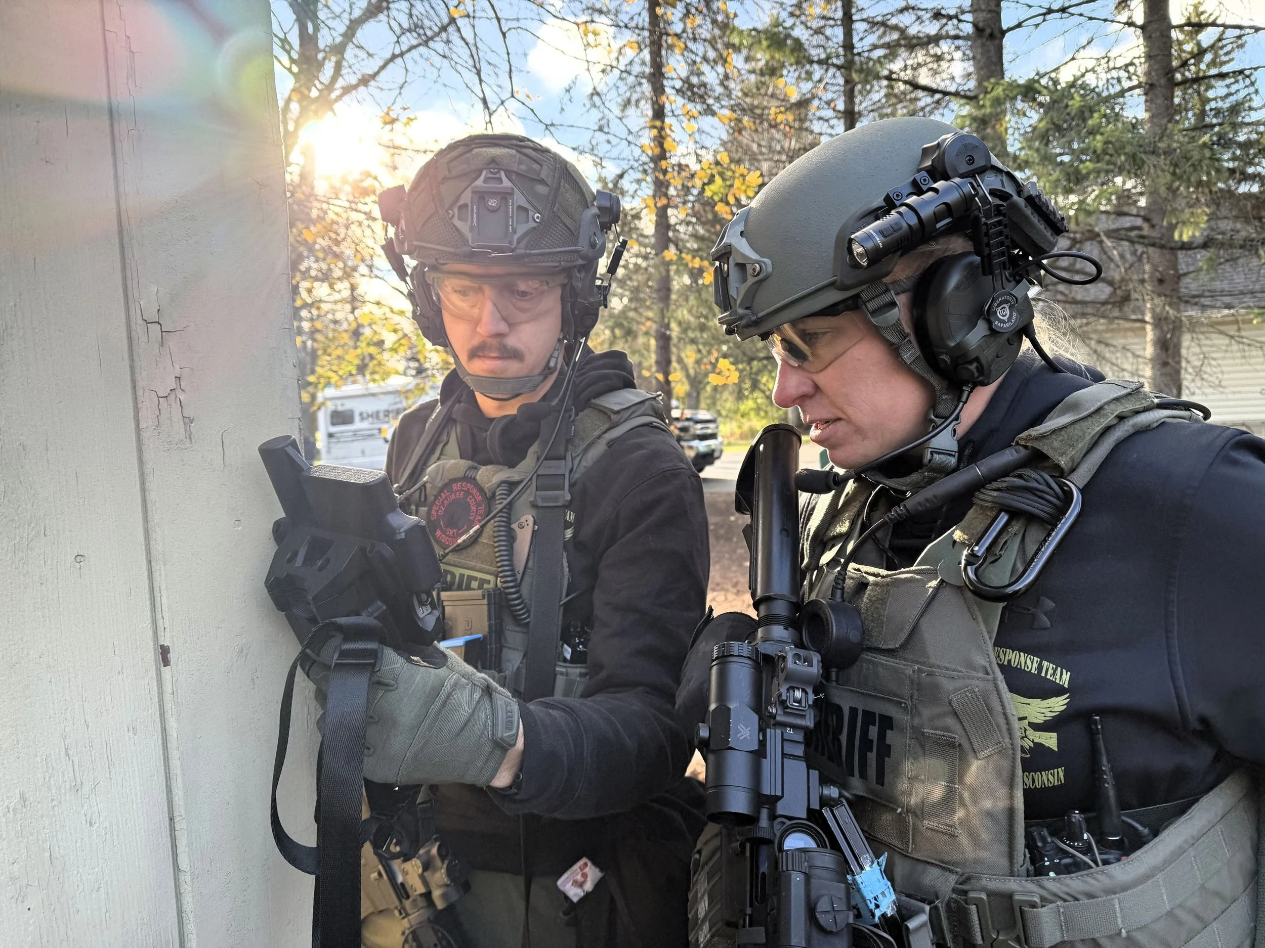 Two police officers wearing tactical gear and helmets, standing outdoors near a wall during daytime. One officer is looking at a device, and the other is speaking into a microphone attached to a helmet, with trees and a bright sky in the background.