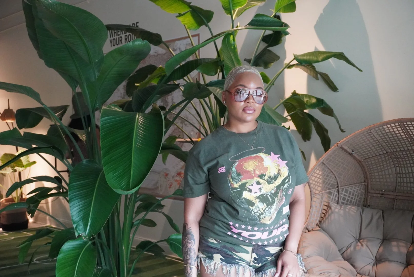 A woman with short blonde hair, wearing sunglasses, a graphic t-shirt with a skull and planet Earth design, and camouflage shorts, standing indoors next to large green houseplants and a wicker seating area.