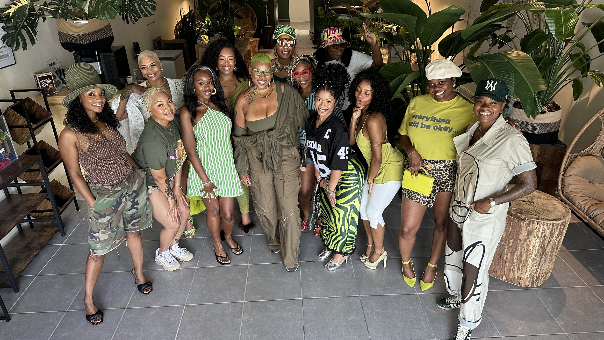 A group of women posing together indoors, surrounded by large green plants and wooden furniture, with some smiling and showing unique fashion styles.