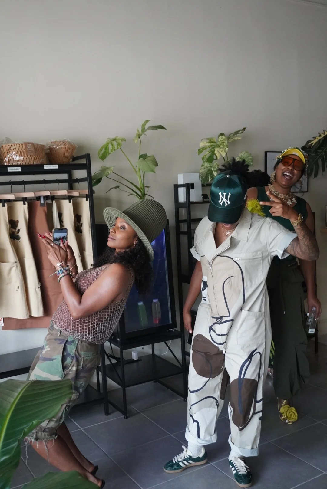 Three women in a room, two wearing hats and one with sunglasses, all appear to be having fun. One woman with a large green hat is looking to the side, another with a black cap and white outfit is dancing or posing, and the third woman in the backgrou